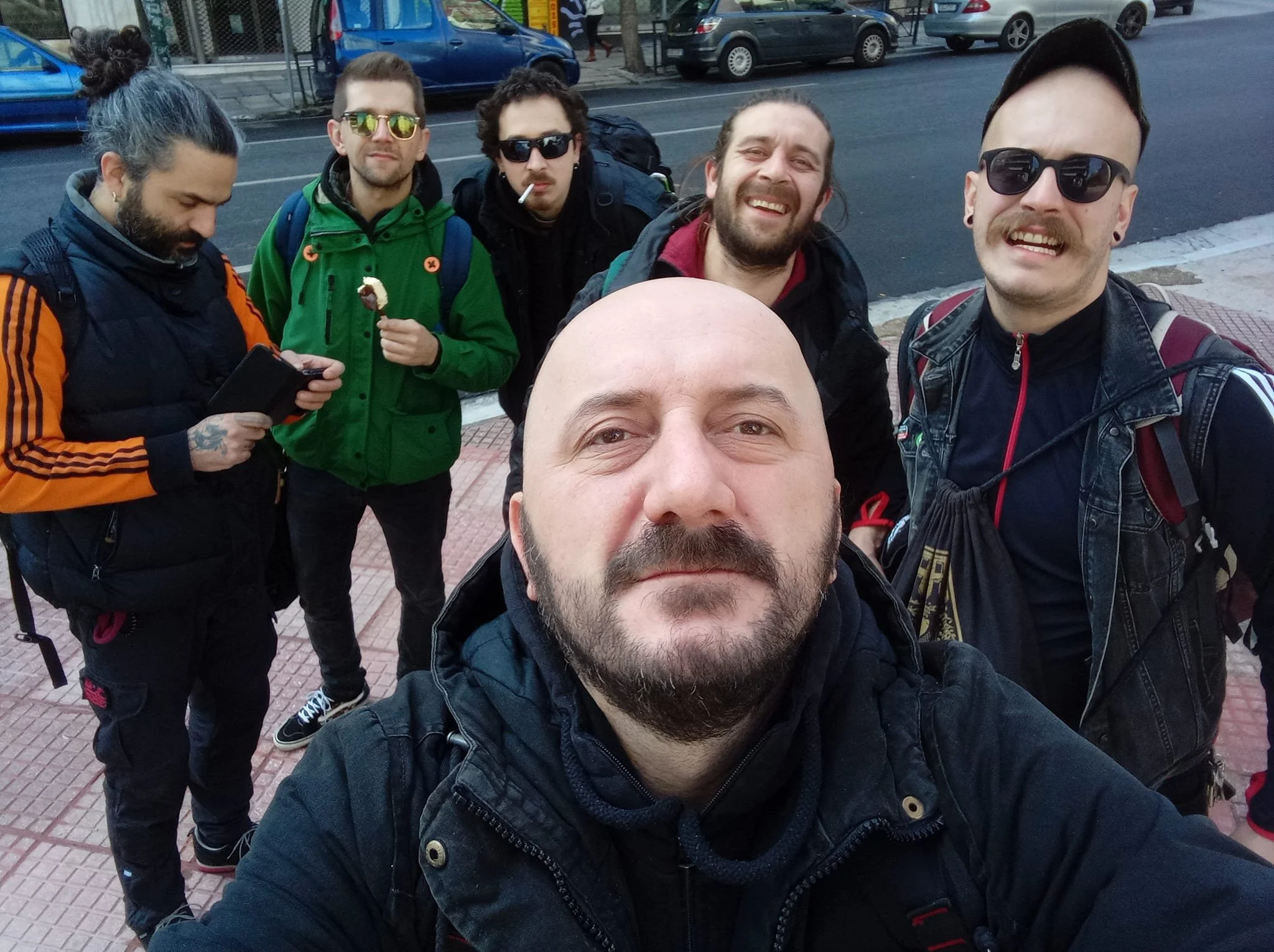 Group of five young men taking a selfie on the sidewalk with cars and trees in the background.