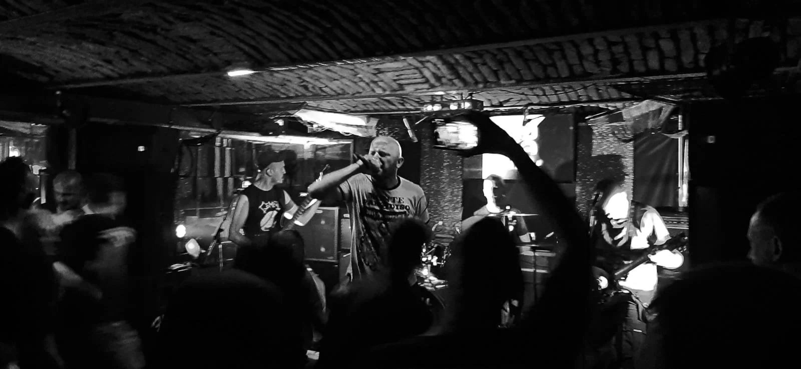 A black and white photo of a live music performance at a bar or club. There is a band on stage with a lead singer holding a microphone, and other band members playing guitars and drums. Audience members are standing close, some raising their hands.