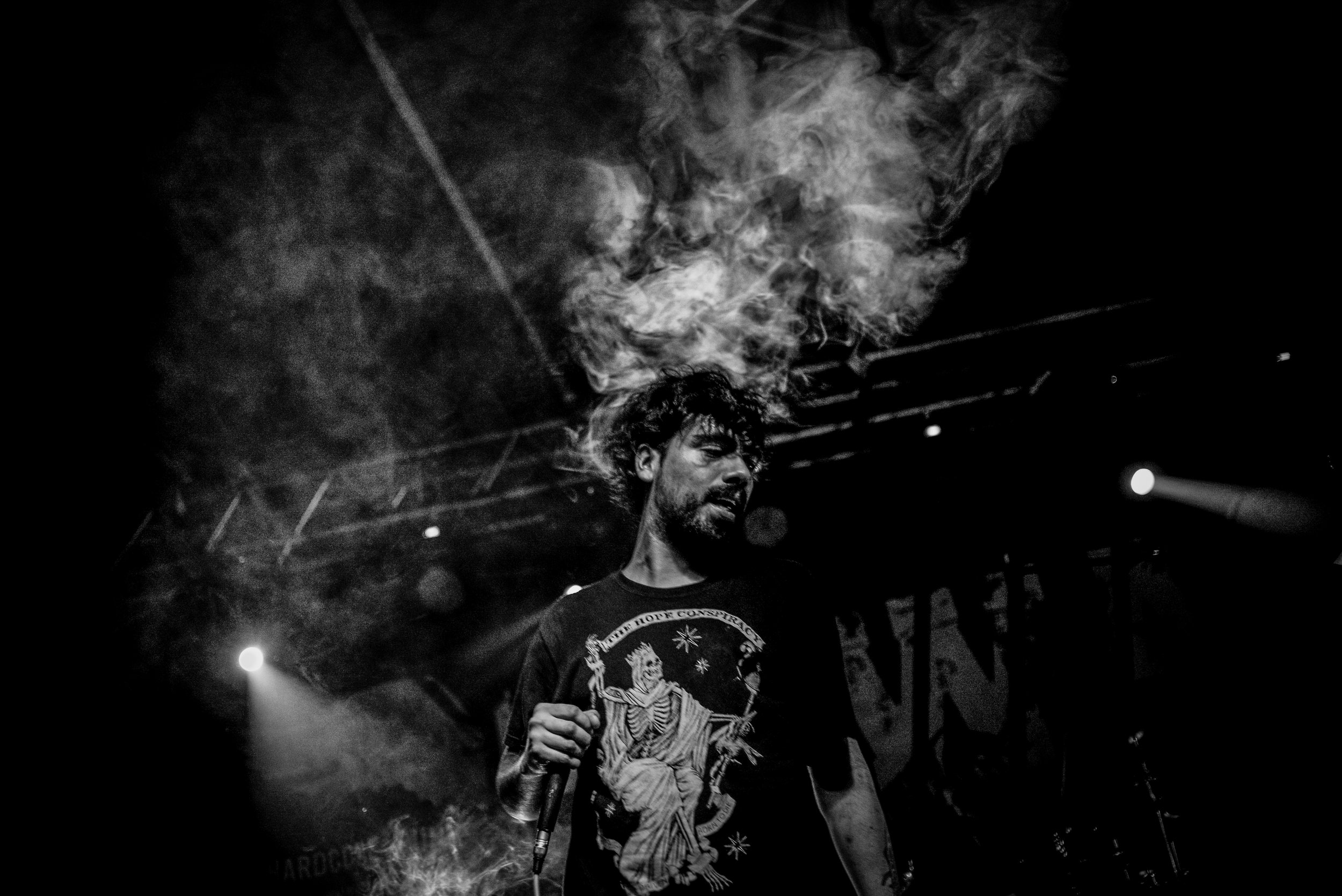 Black and white image of a man with curly hair and tattoos, holding a microphone, with smoke and stage lights in the background. Band name: En Mi Defensa