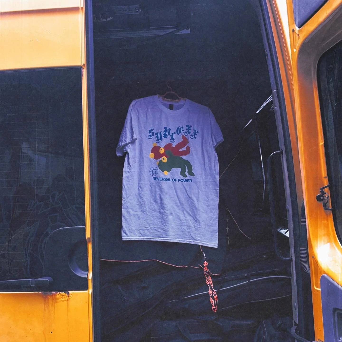 White T-shirt with graphic design hanging on a hanger inside a glass display case, with a black background. The graphic features a colorful creature with pentagram symbols and text that reads 'Reversal of Power'.