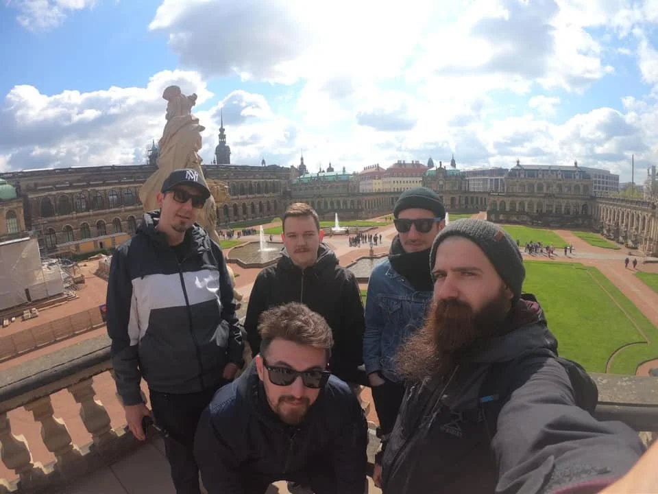 Five men posing for a selfie on a balcony overlooking a historic European palace with statues, fountains, and a large green lawn, under a partly cloudy sky.