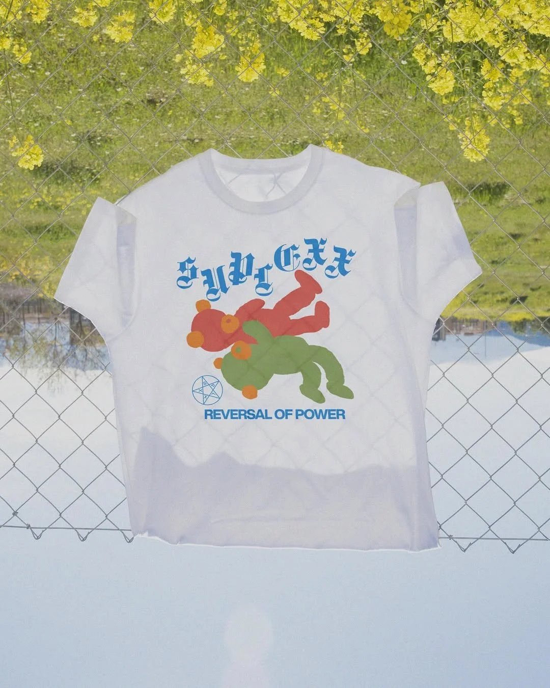 White graphic T-shirt featuring cartoonish red and green robots with orange accents, surrounded by blue text in a stylized font, a blue pentagram symbol, and the phrase "REVERSAL OF POWER". The T-shirt is hanging outdoors against a chain-link fence w
