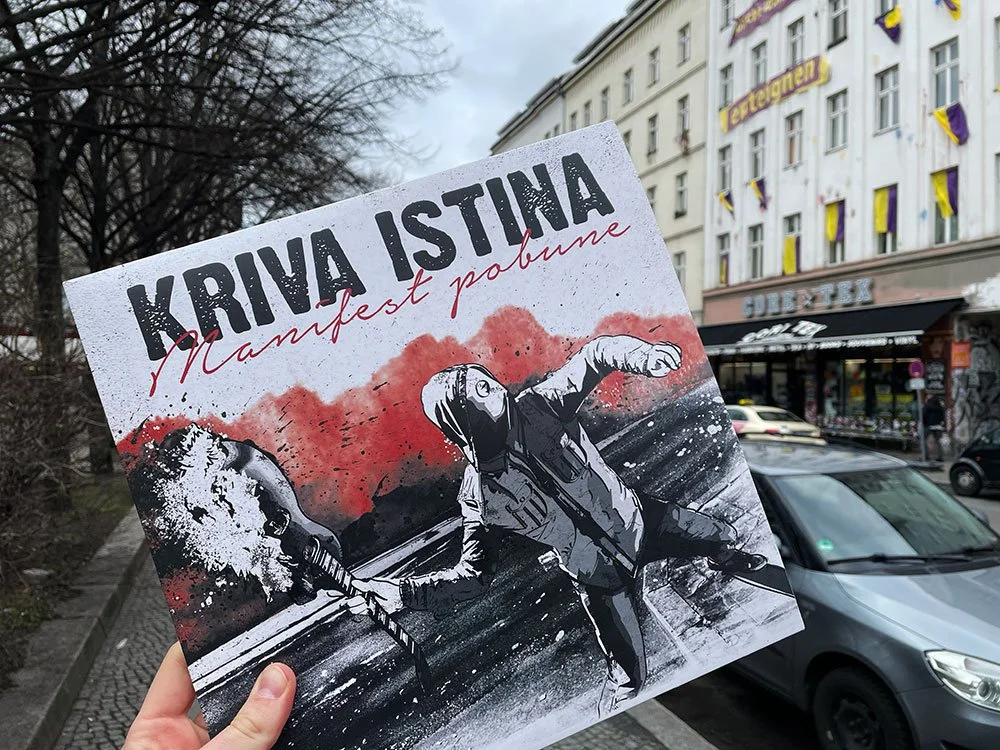 A person holding a protest sign in front of a city street with cars and buildings. The sign features a black and white illustration of a person holding a flag or torch, with bold black and red text that reads "KRIVA ISTINA" and "Manifest Pobune".