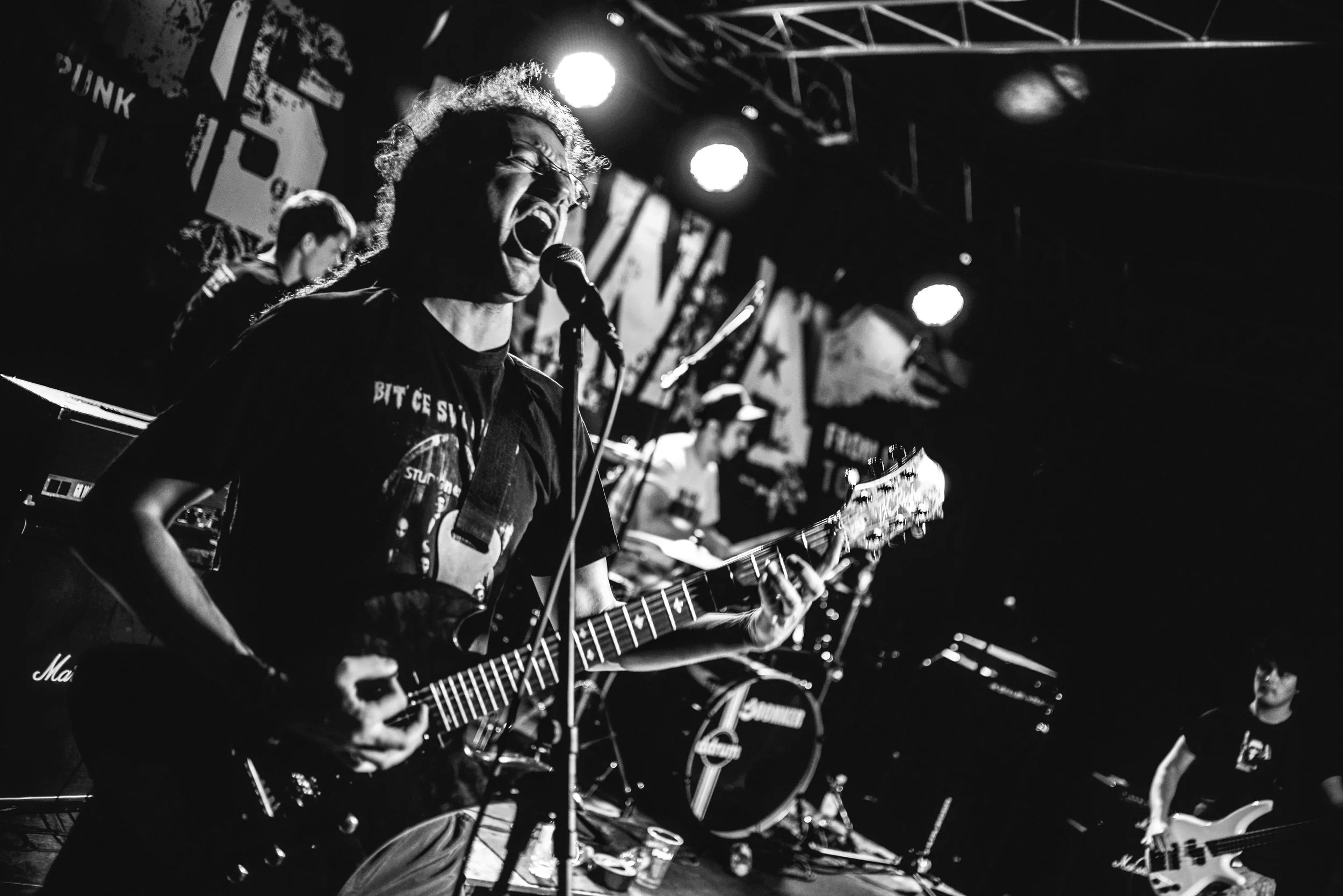 Black and white photo of a musician passionately singing and playing an electric guitar on stage, with a microphone and stage lights in the background.