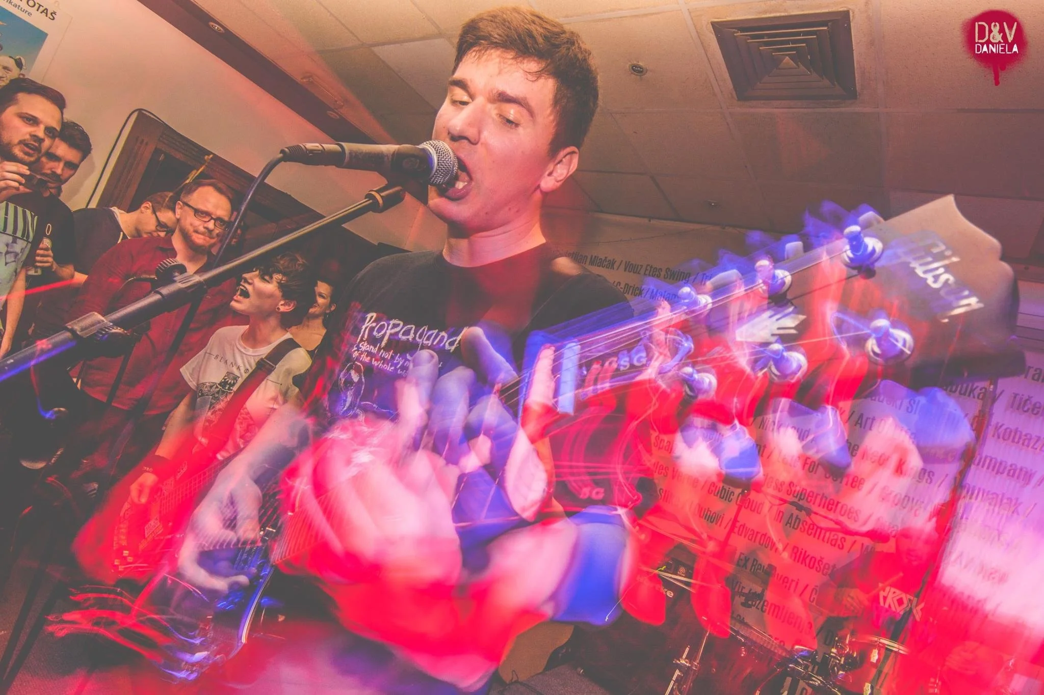 A person singing into a microphone while playing a guitar, with a group of people in the background at an indoor event.