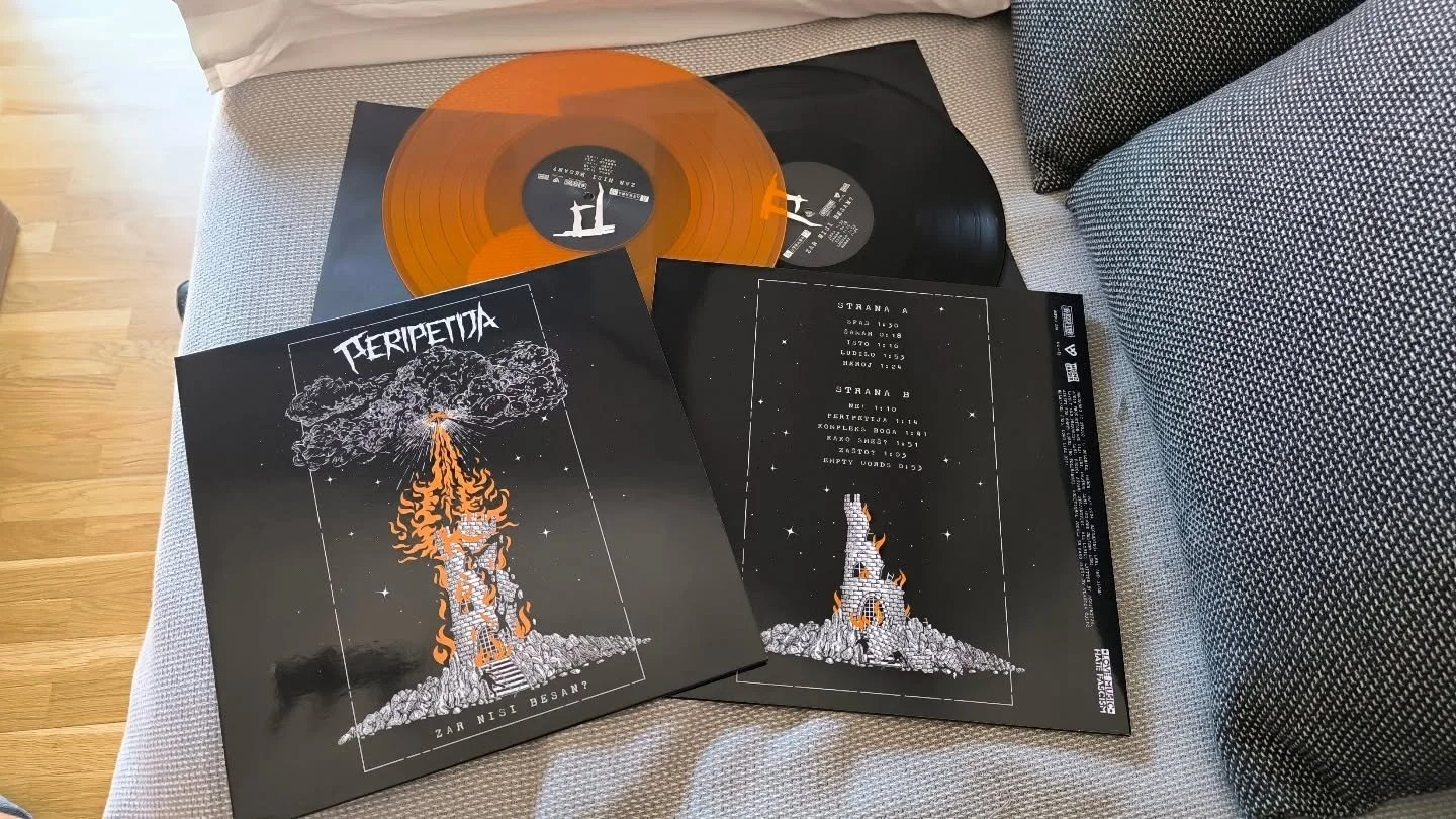 Two vinyl records and two album covers on a gray fabric surface. The records are black and orange, with one partially overlapping the other. The album covers are black with white and orange artwork; one features a flaming tower with smoke, and the ot