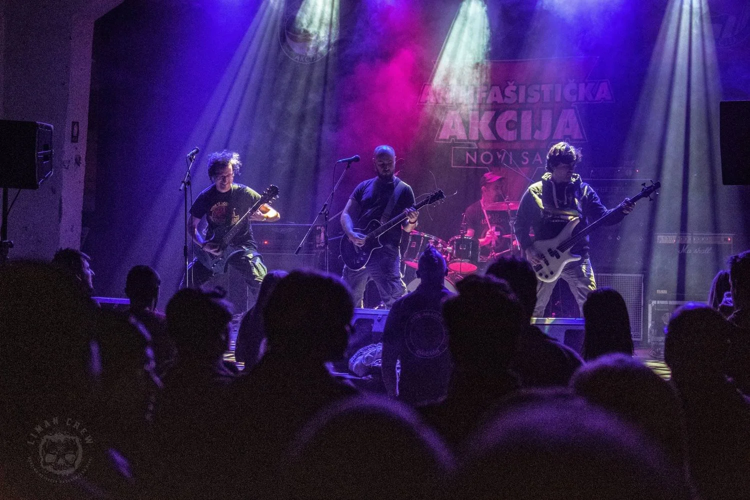 A band performing on stage at a concert with purple lighting. The band consists of four members playing guitars and drums, with an audience watching in the foreground. There is a banner with text in the background.