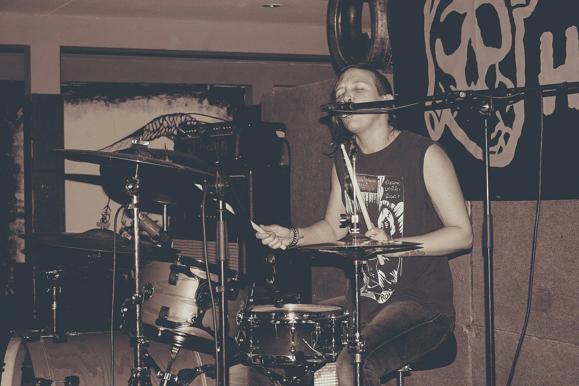 A woman with short dark hair playing drums and singing into a microphone at a live music event. She is wearing a black sleeveless shirt and appears to be passionately engaged in her performance.