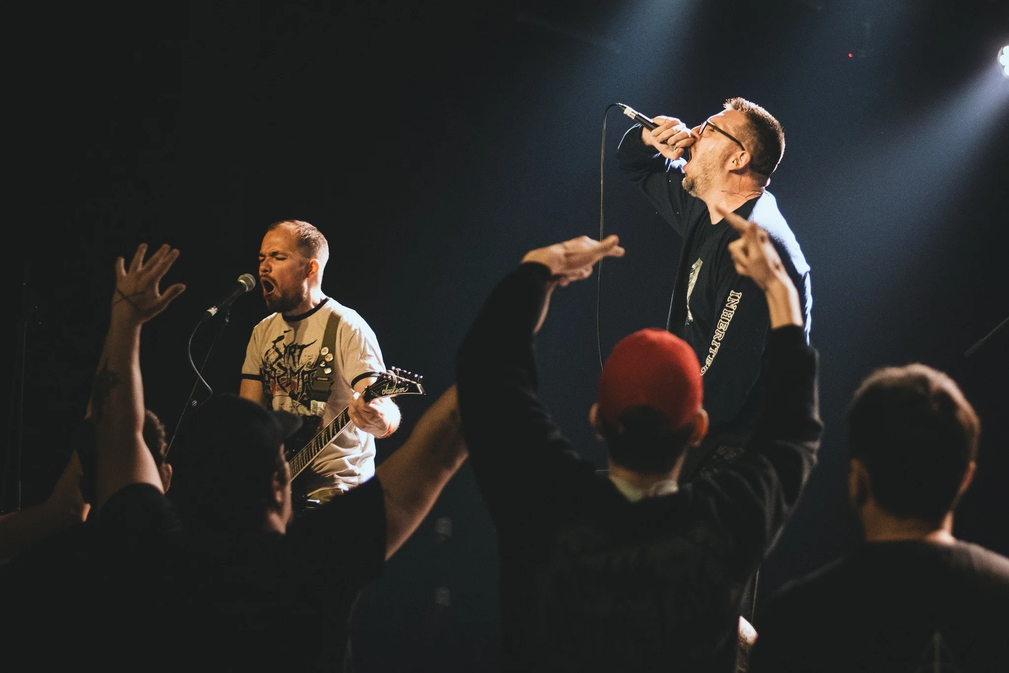 Two male performers on stage, one singing into a microphone with eyes closed and guitar in hand, the other passionately singing or yelling into a microphone with glasses on, surrounded by an enthusiastic crowd with raised hands.
