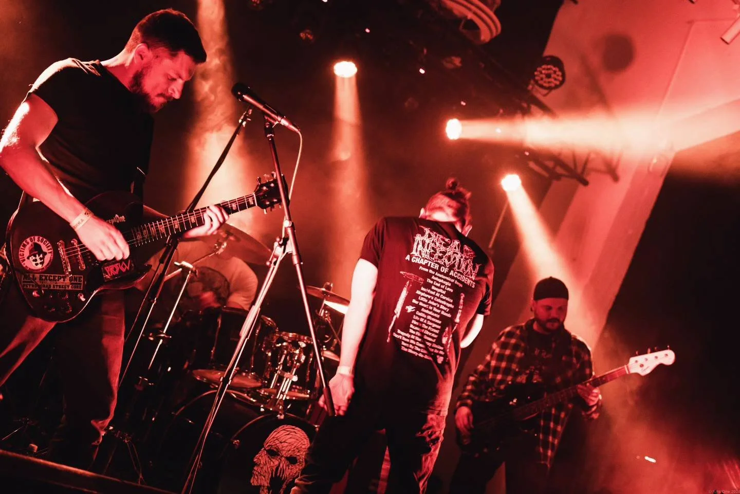 A band performs on stage with red lighting, featuring a guitarist, a bassist, and a drummer, with haze and stage lights creating a vibrant atmosphere.