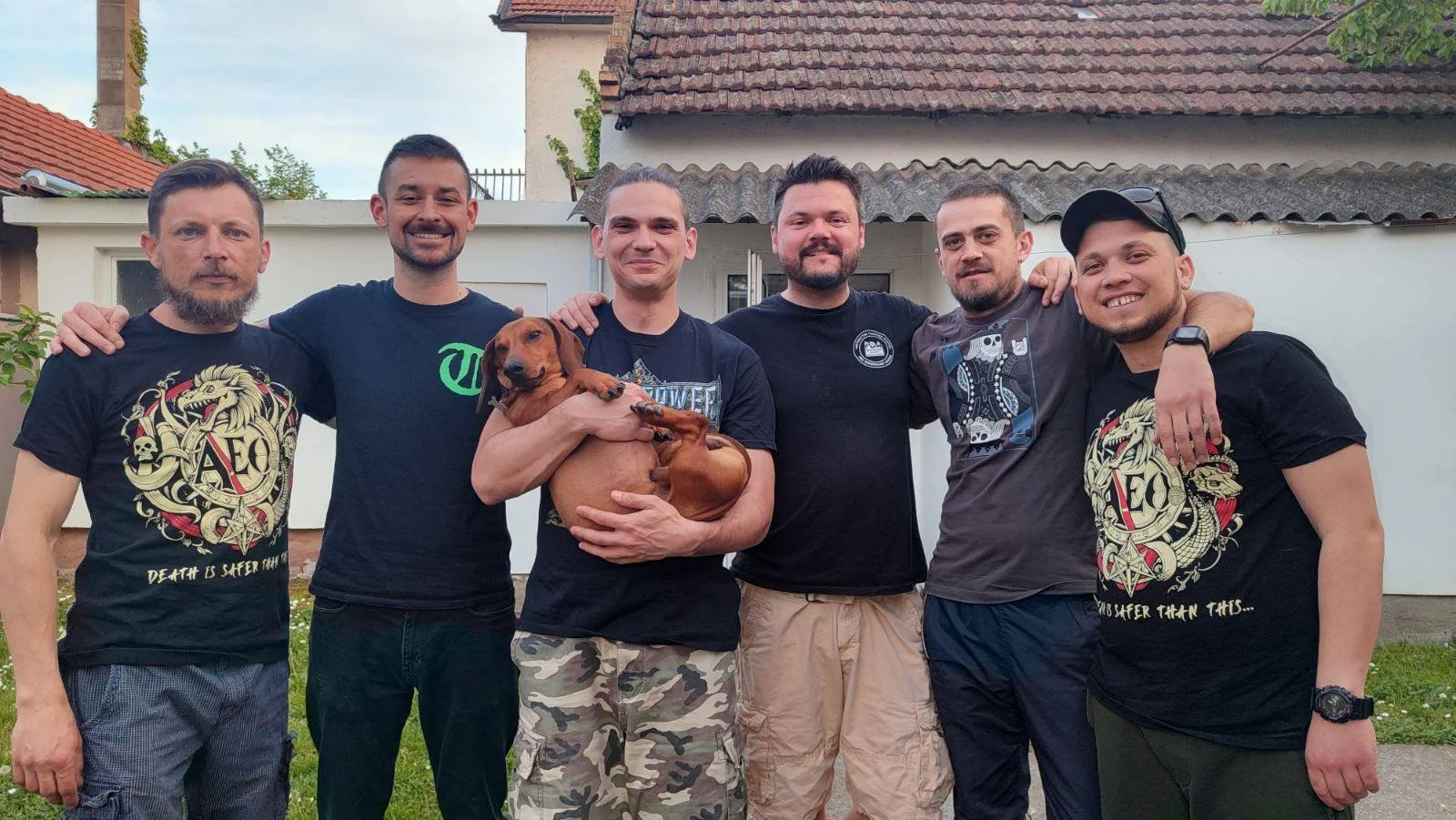 A group of six men standing outdoors in a backyard, some with their arms around each other, with a man in the middle holding a brown dog. They are smiling and dressed casually, with houses and a white fence in the background.