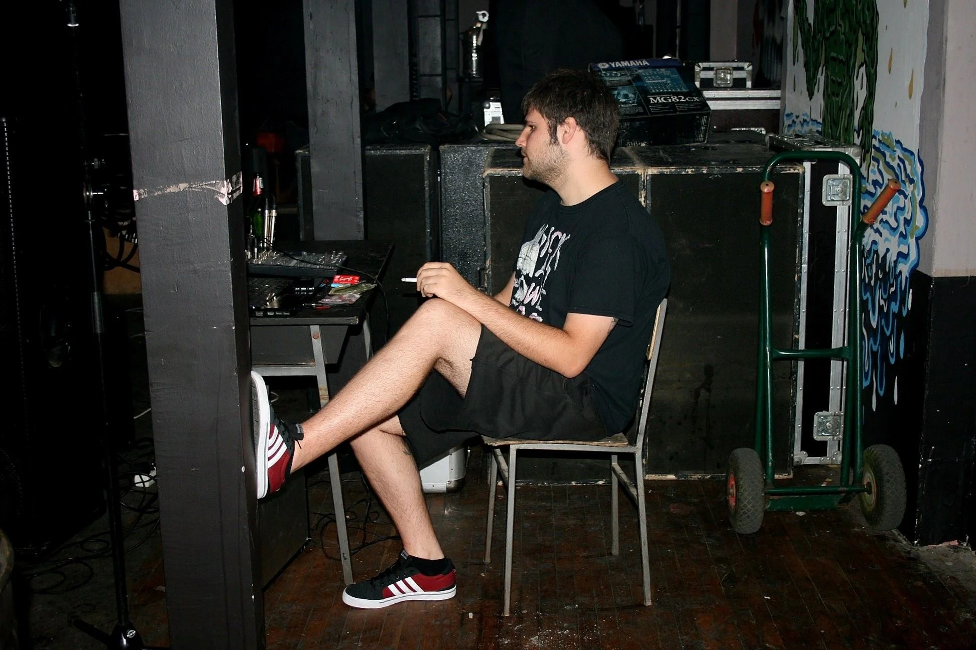 A young man with dark hair and a beard sitting in a backstage area, using a laptop. He is wearing a black T-shirt and striped sneakers, with his legs crossed and resting on the table. There are various equipment and tools around, and a green hand tru
