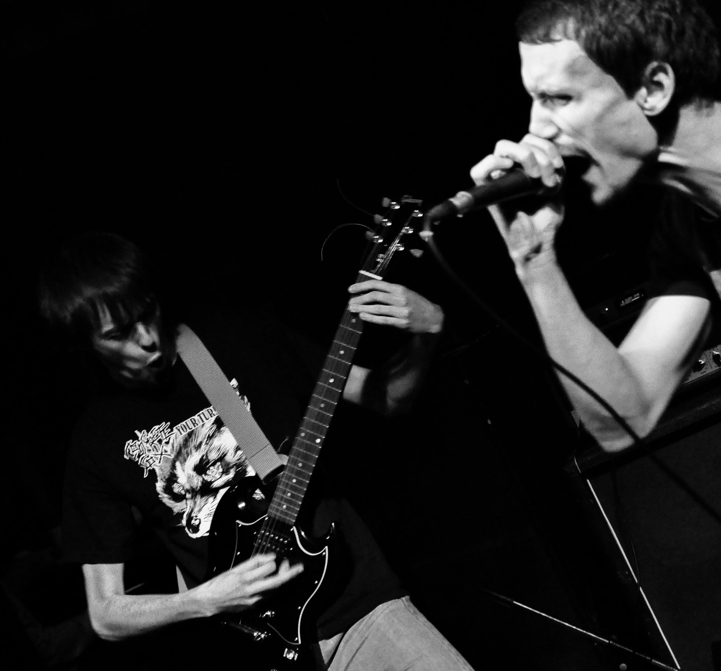A black-and-white image showing a young person singing into a microphone while playing an electric guitar. They are on stage, and their facial expression shows concentration or emotion.