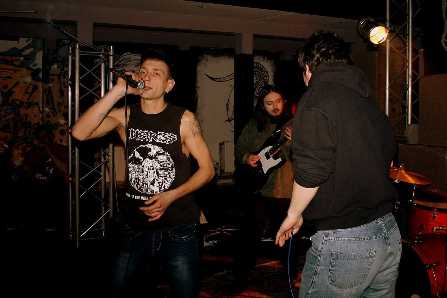 Band performing live on stage with singer holding microphone, guitarist playing guitar, and drummer visible in background.