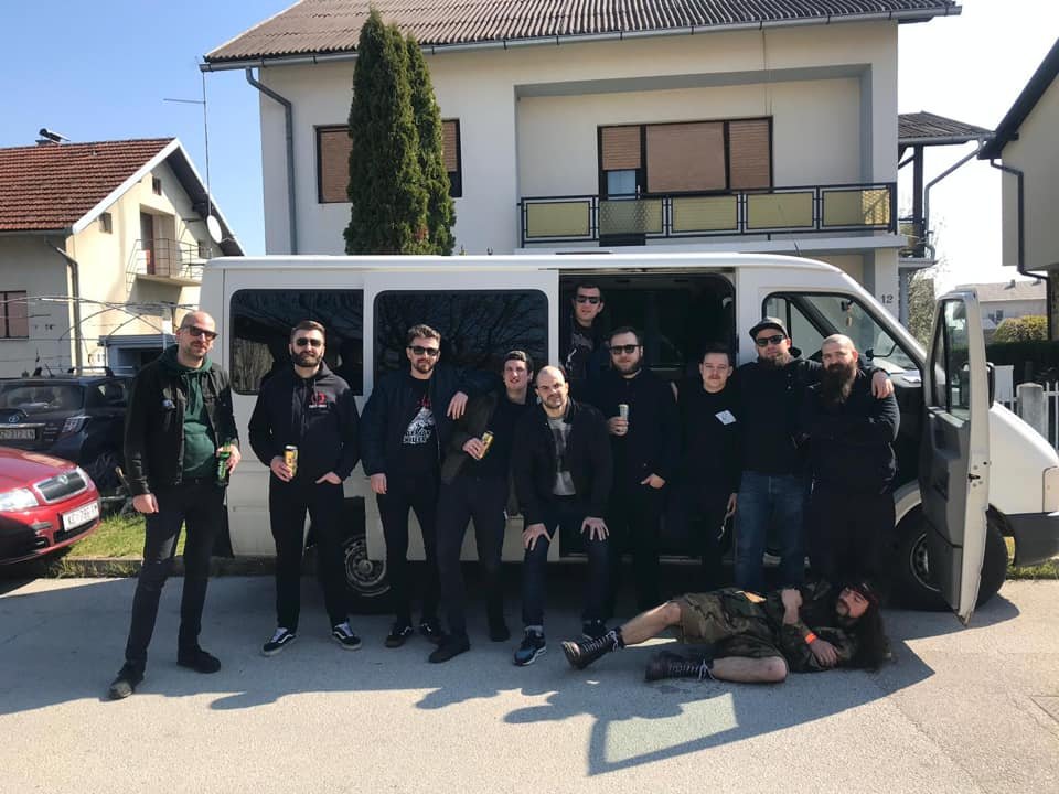 Group of men standing in front of a white van, some holding drinks, with one person lying on the ground in front of the van, all outside in a residential neighborhood.