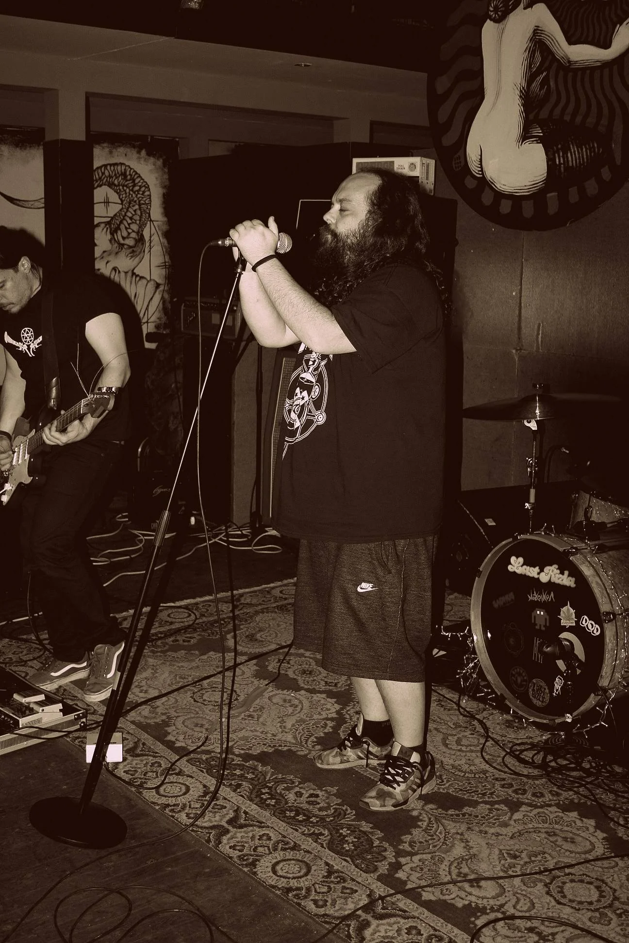 A man with long hair and a beard singing into a microphone on stage with a guitar player beside him. There are musical equipment including drums and amplifiers in the background.
