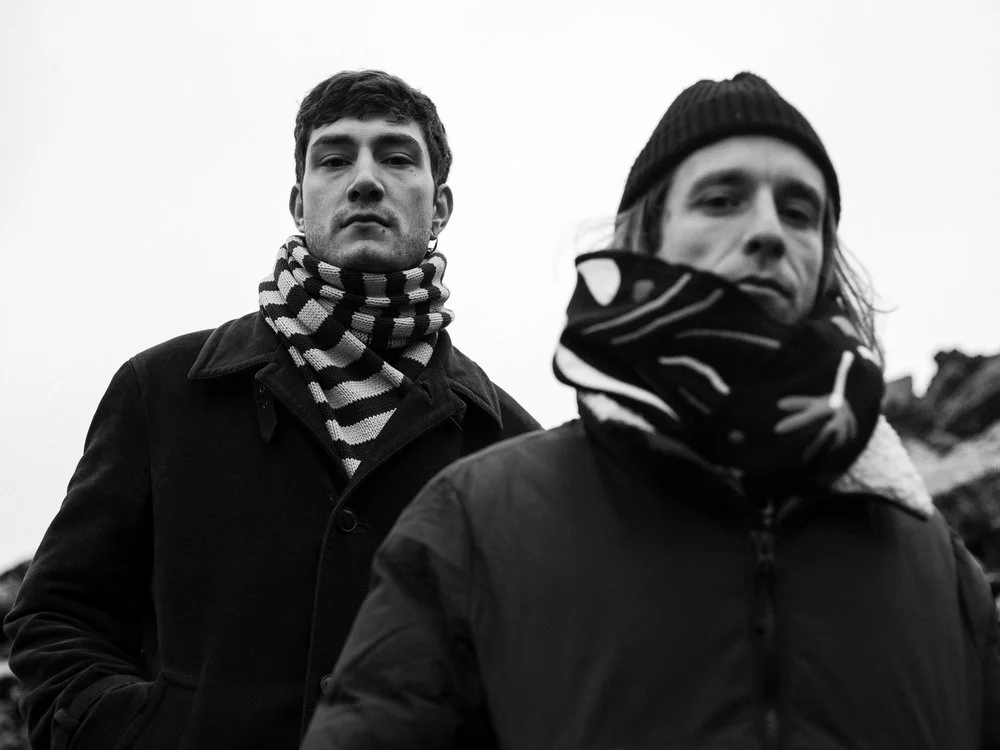 Two men outdoors in winter clothing, one in a black coat and striped scarf, the other in a beanie and patterned scarf, with a plain sky background.