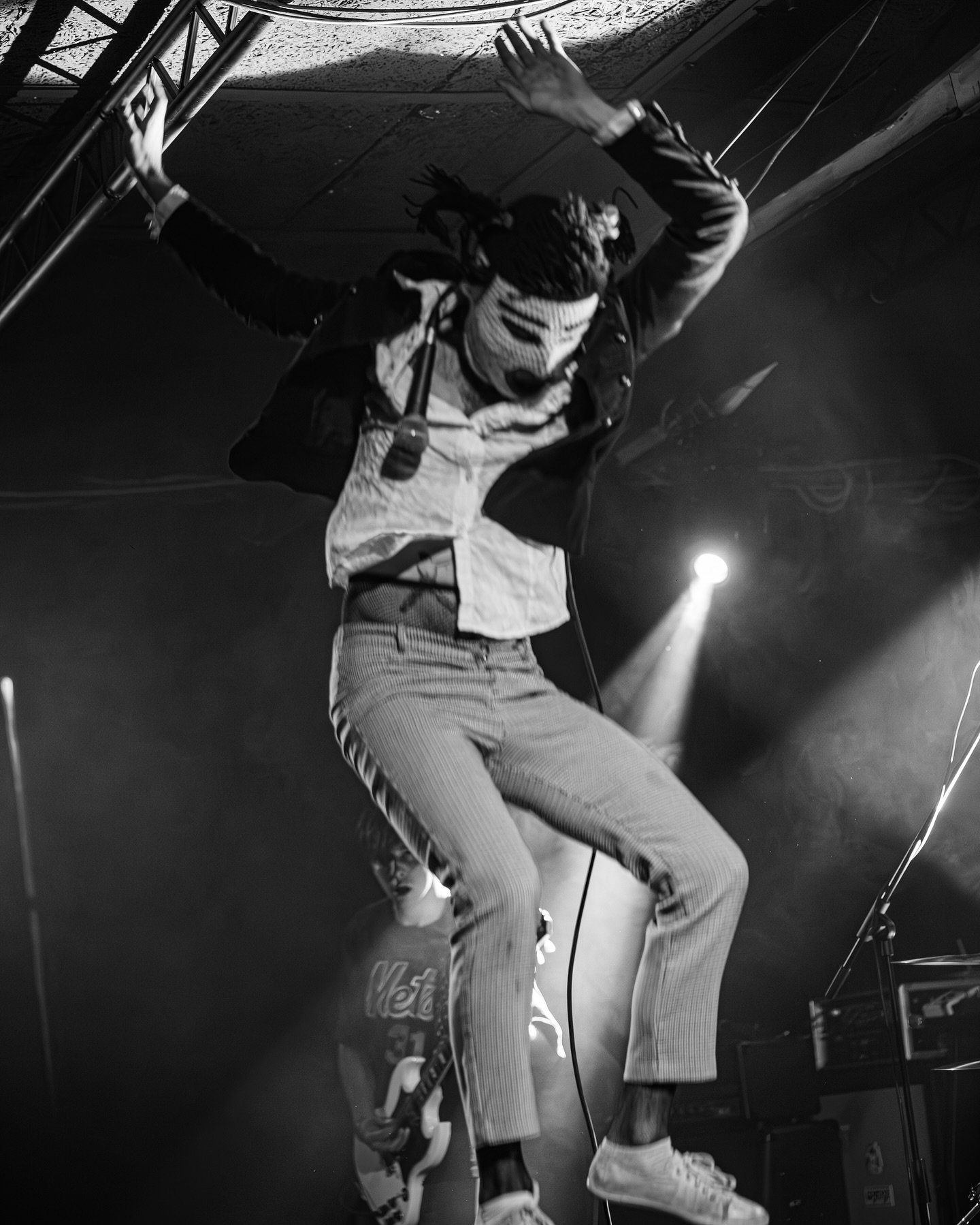 A person with dreadlocks and a white face mask performing a dance move on stage. They are wearing a jacket, striped pants, and white sneakers. The stage has a metal scaffolding and stage lighting.