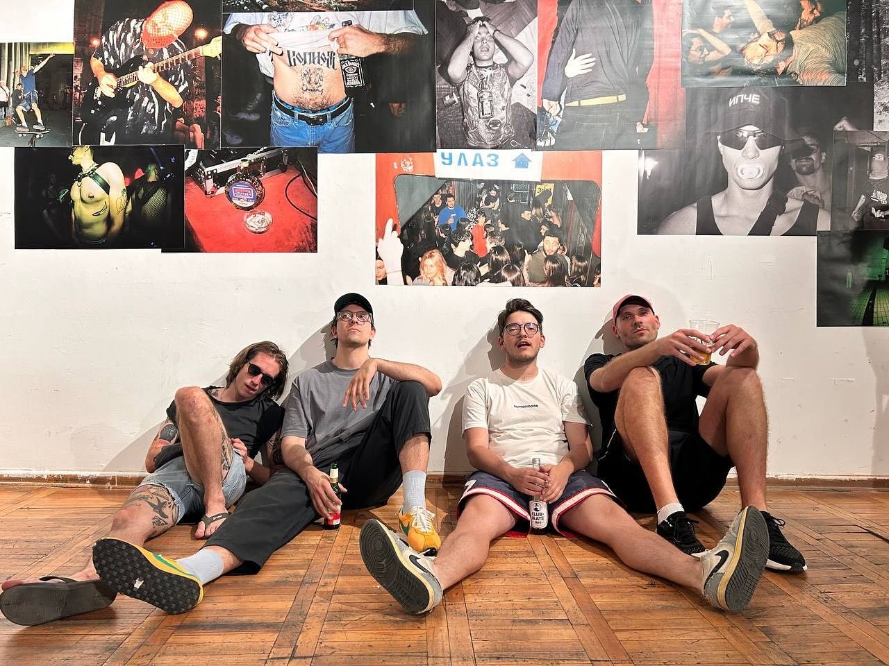 Four young men sit on the wooden floor against a white wall decorated with various photos of people at a party or concert. The men are casually dressed, some with drinks in hand, and appear relaxed.