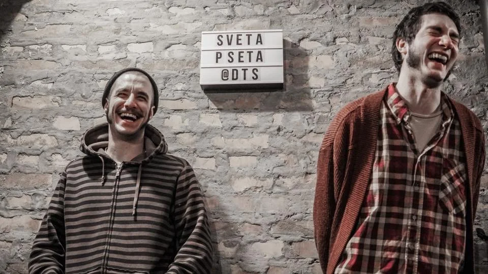 Two men laughing and standing against a brick wall with a lightbox sign that reads 'SVETA PSETA' above their heads.