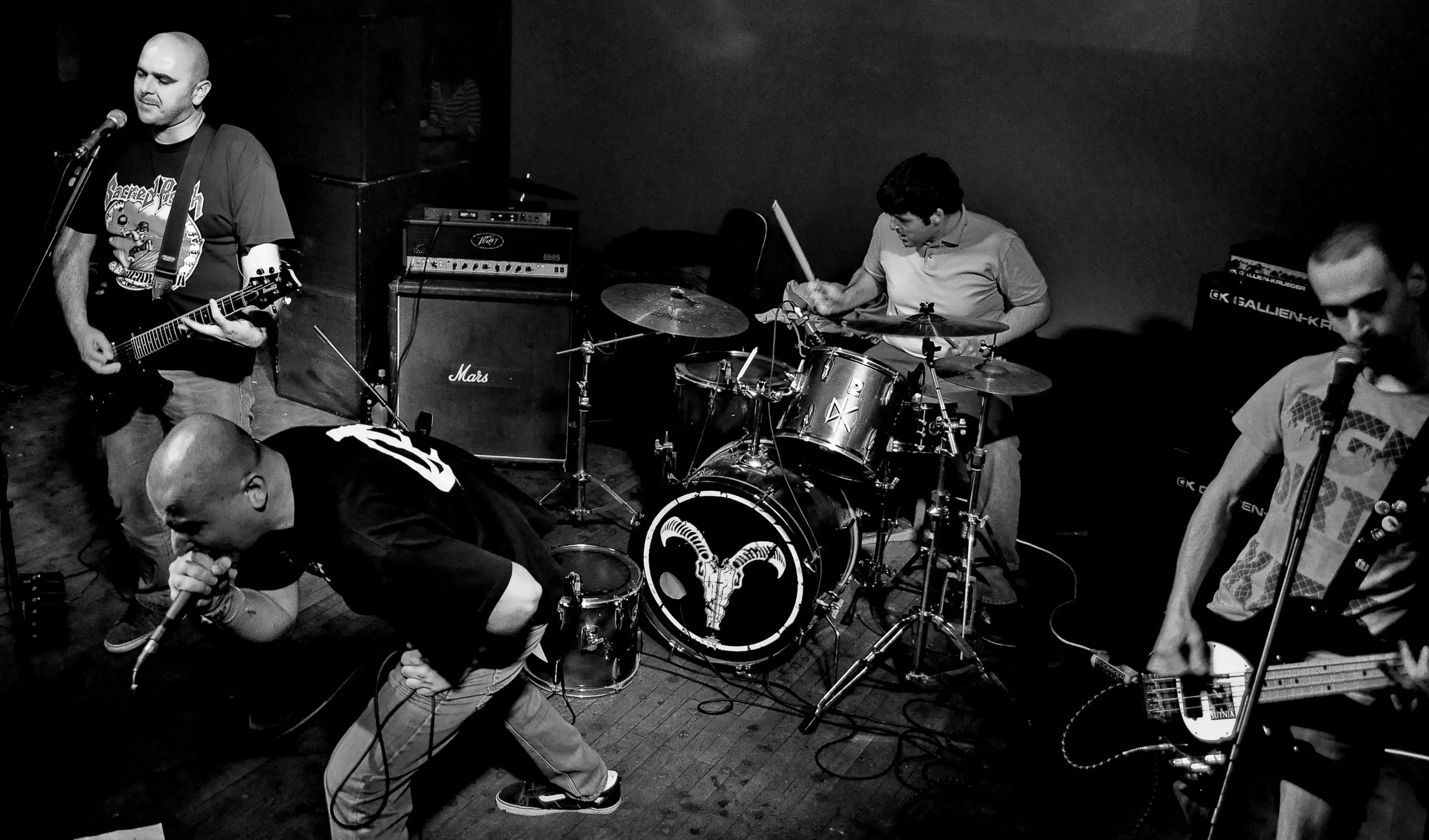 A live performance of a rock band on stage with five members. One is singing into a microphone and playing an electric guitar, another is crouching on the floor singing with a microphone, the third is playing the drums, and two others are playing gui