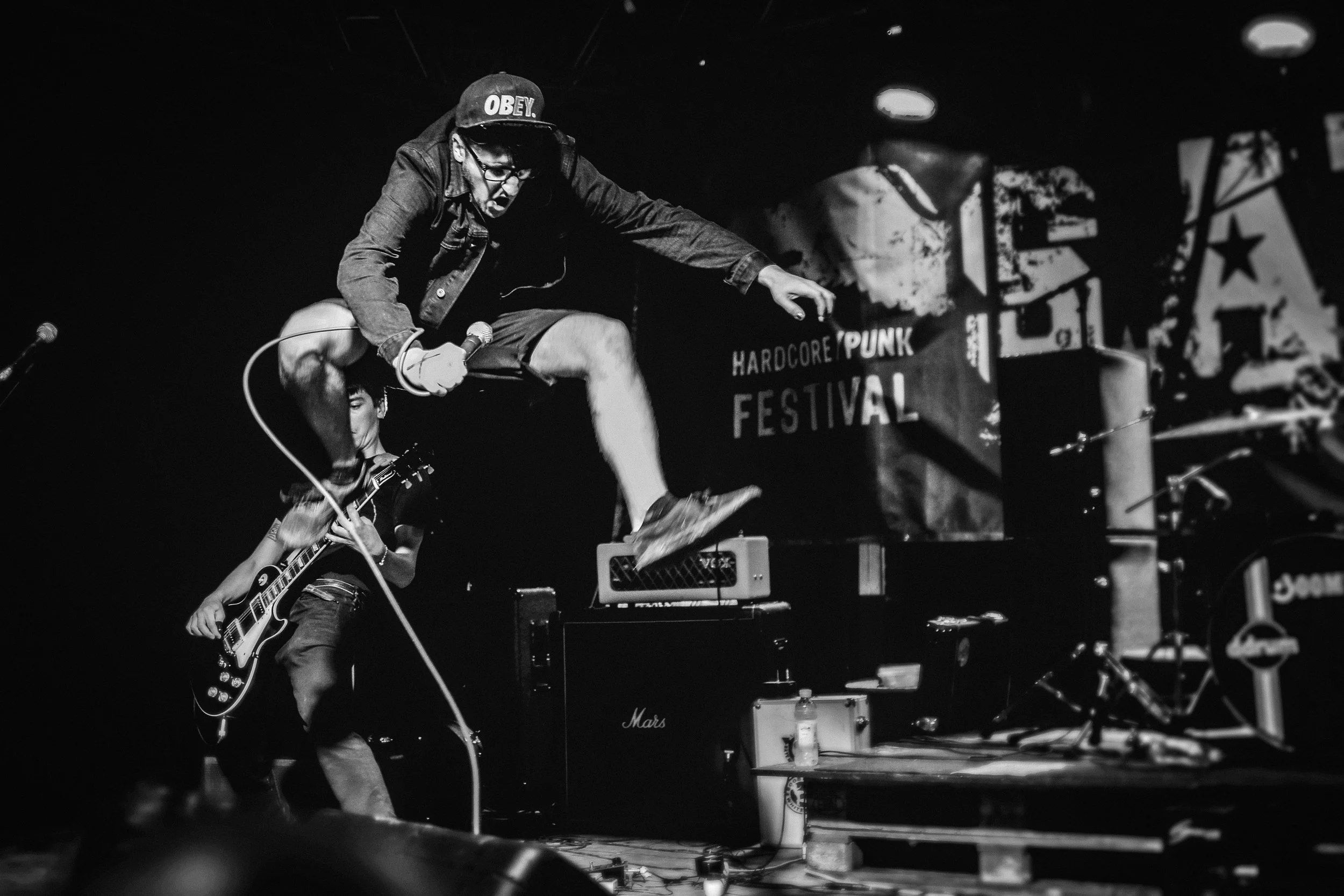A black and white photo of a punk rock band performing live on stage. The lead singer, wearing an 'OBEY' baseball cap and glasses, is jumping or falling forward while holding a microphone. The guitarist, slightly behind, is playing an electric guitar