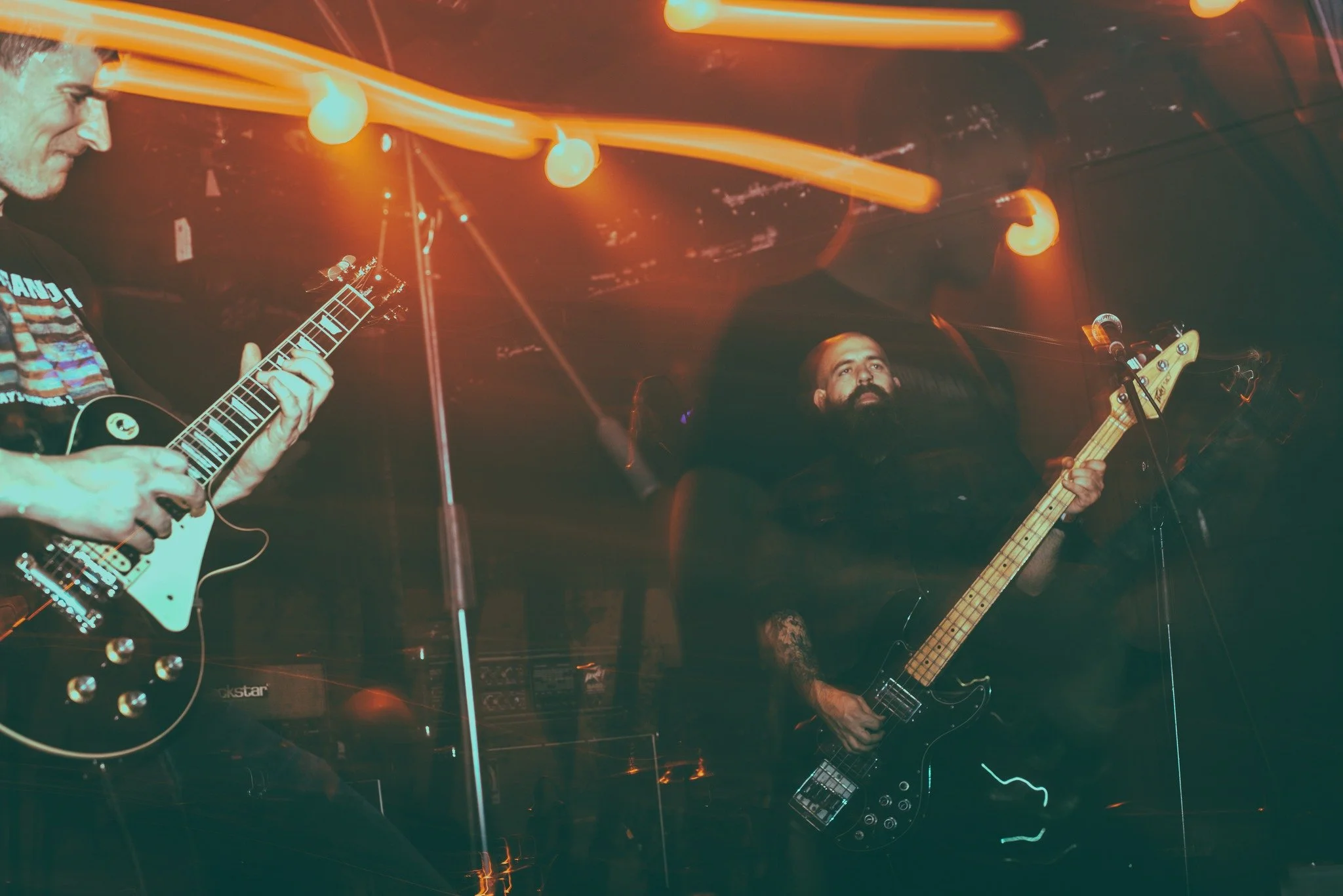 Two musicians performing on stage with guitars under orange stage lighting, one with a black electric guitar and the other with a bass guitar, in a dimly lit venue.