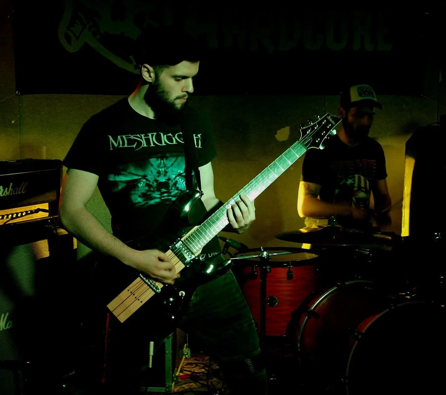 Two musicians performing in a dimly lit room. One plays an electric guitar, and the other plays drums. Both wear dark T-shirts, and there's audio equipment visible.