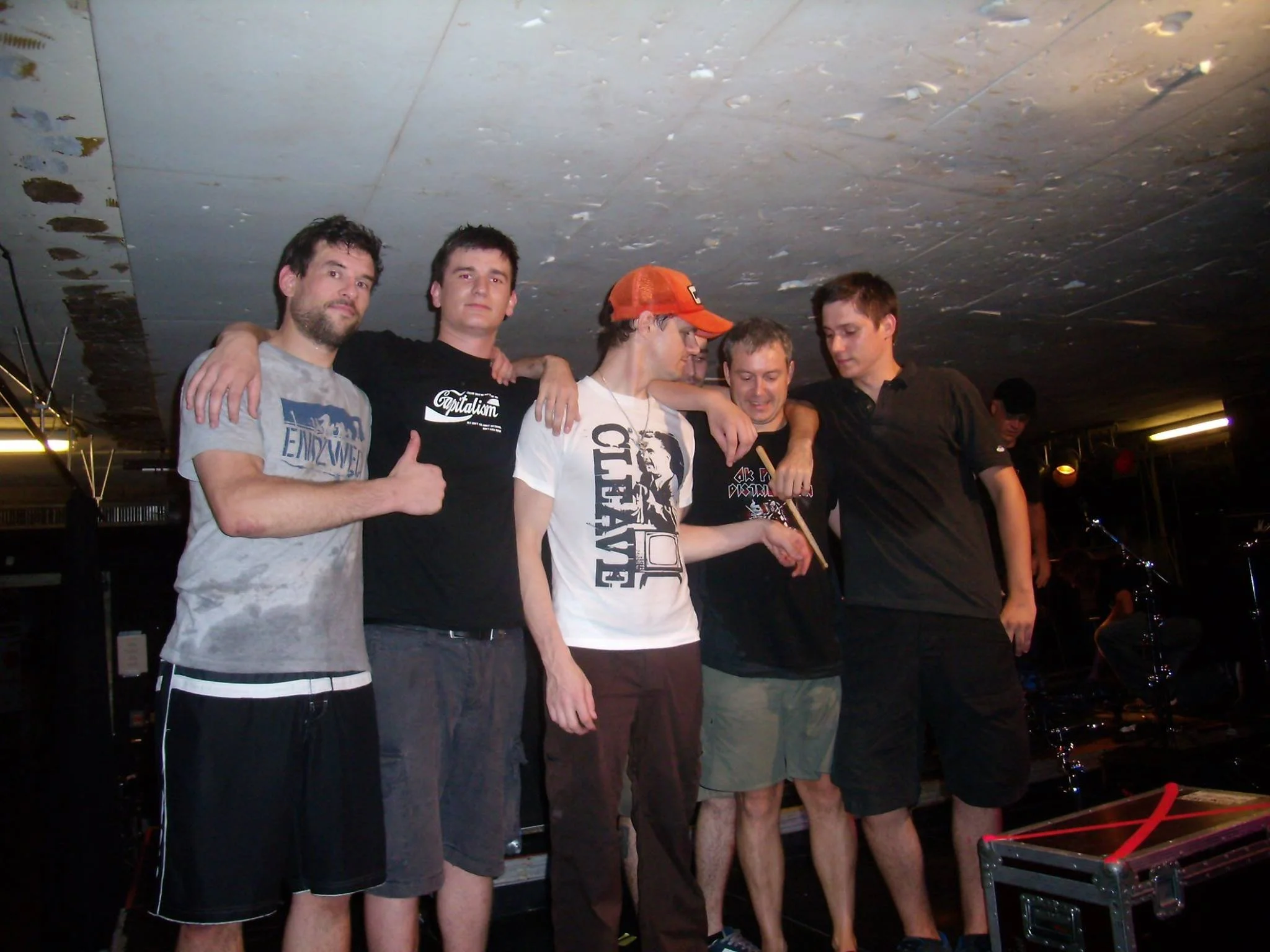 A group of five young men standing closely together with their arms around each other, smiling, in an indoor setting with a black ceiling and some equipment visible.