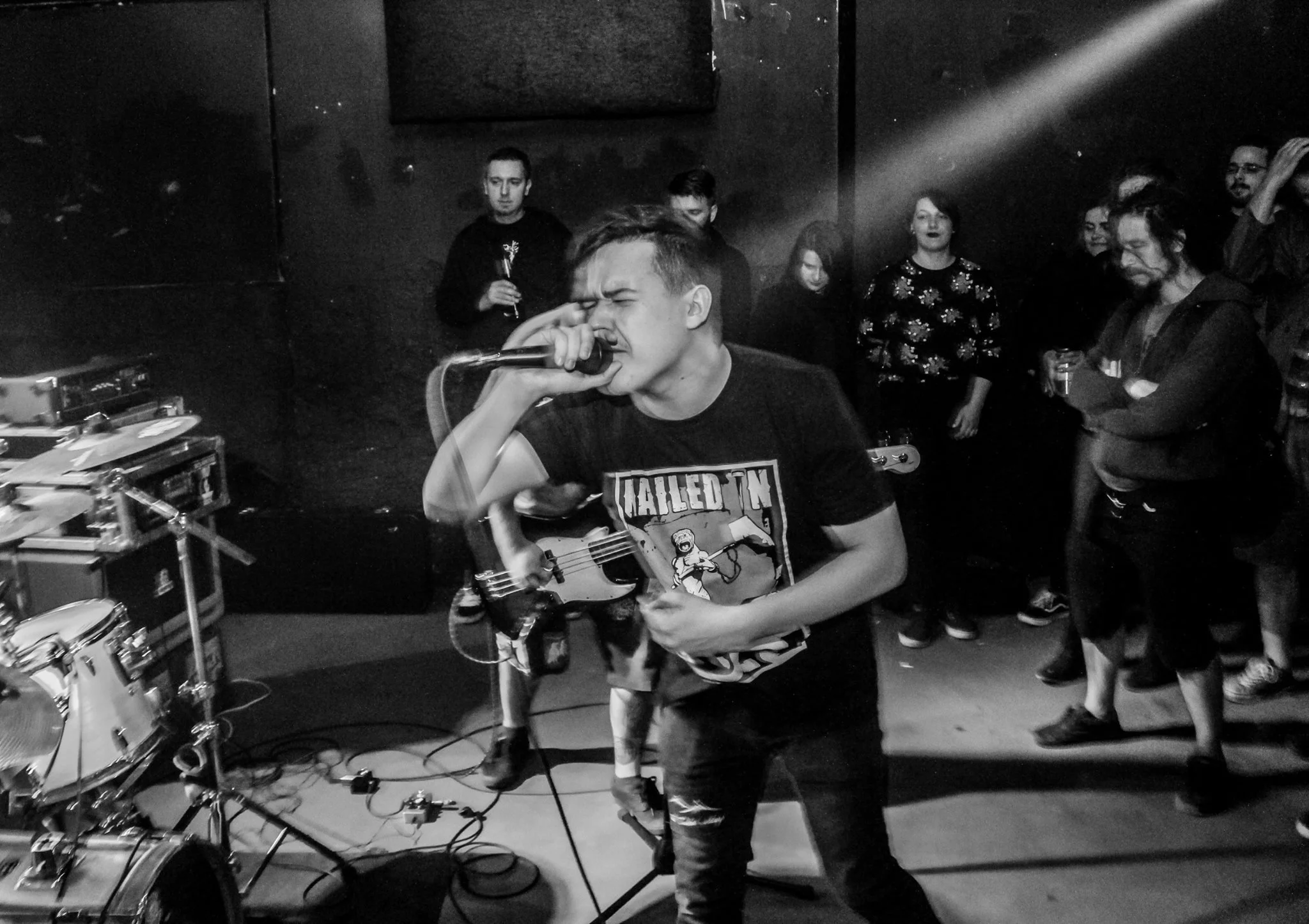 A young man singing with a microphone during a live music performance surrounded by a small audience in a dark indoor venue.