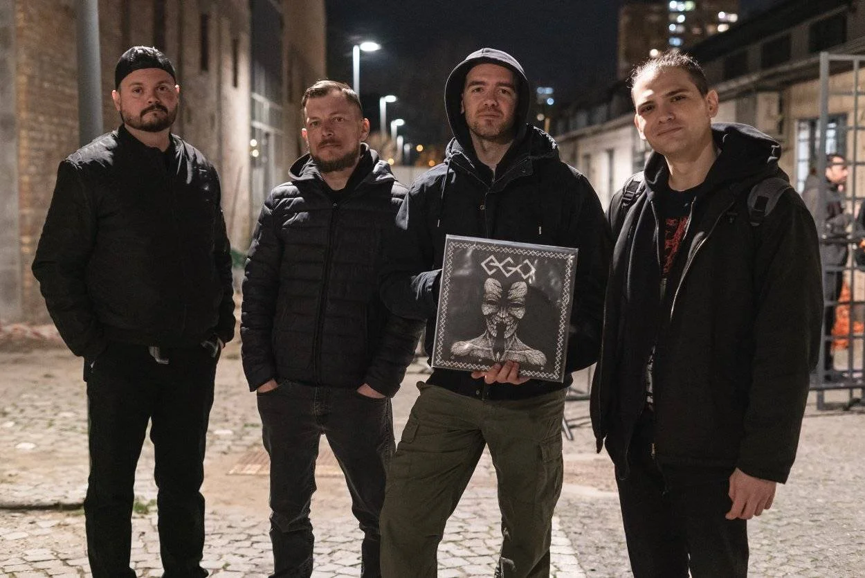 Four men standing on a city street at night, dressed in dark jackets. The man second from the right holds a vinyl record with a black-and-white artwork of a face with a mask and a butterfly overlay.