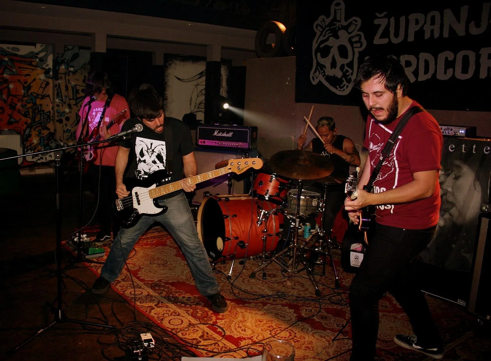 A band performing on stage with three musicians clearly visible, playing guitars and drums. The stage has dark walls with abstract artwork and a Marshall amplifier in the background.