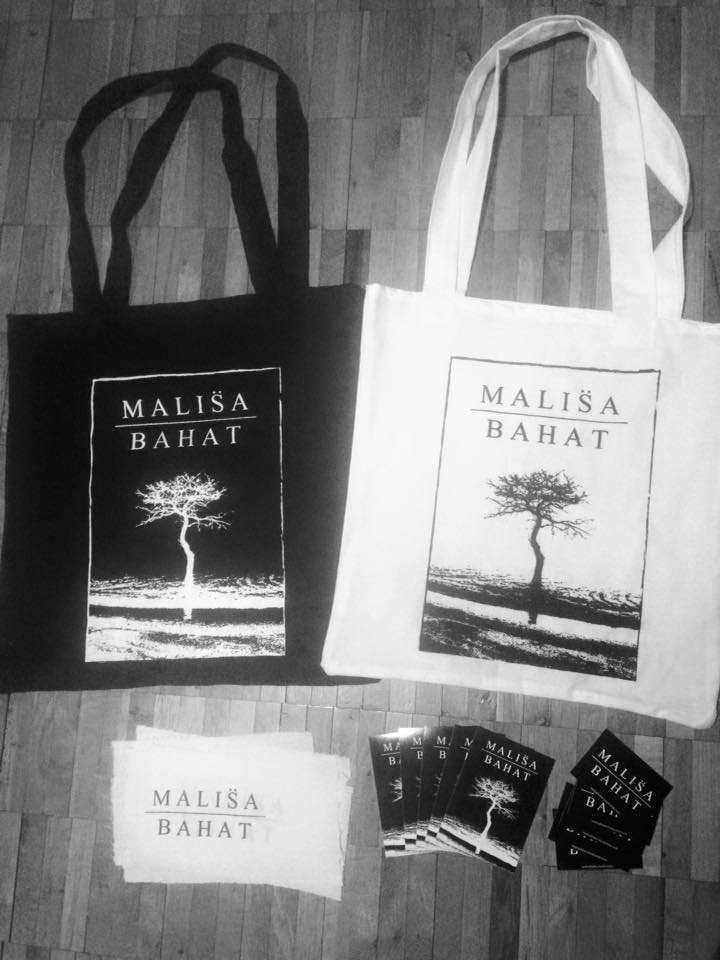 Two tote bags, one black and one white, with a tree graphic and the text 'MALIŠA BAHAT' printed on them. Several smaller cards or booklets with the same design are also displayed on a wooden surface.