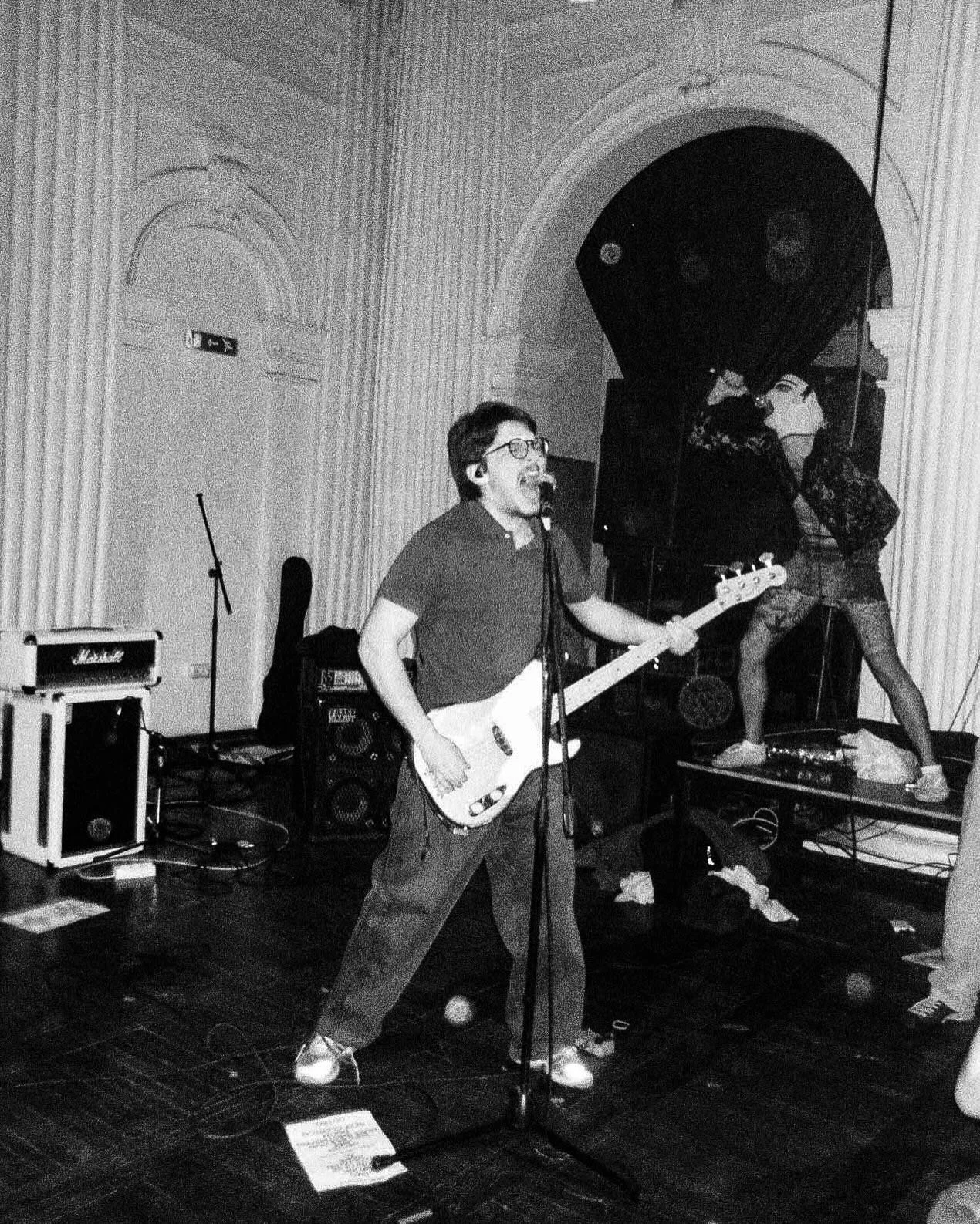 A person singing and playing an electric guitar on stage, with a band member in the background, surrounded by musical equipment in a room with ornate architecture.