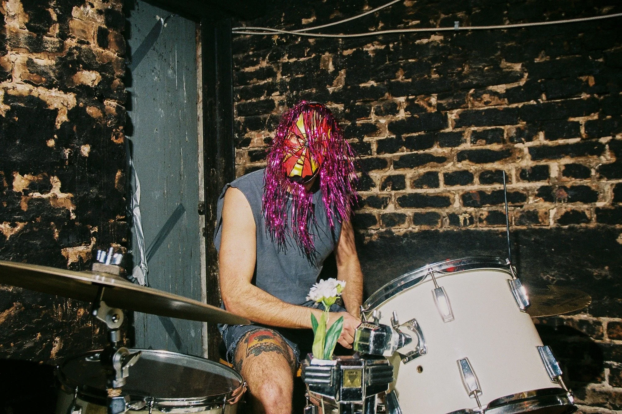 A person with colorful tinsel hair wearing a mask, shirtless, playing a large white drum set in front of a brick wall in a dimly lit room.