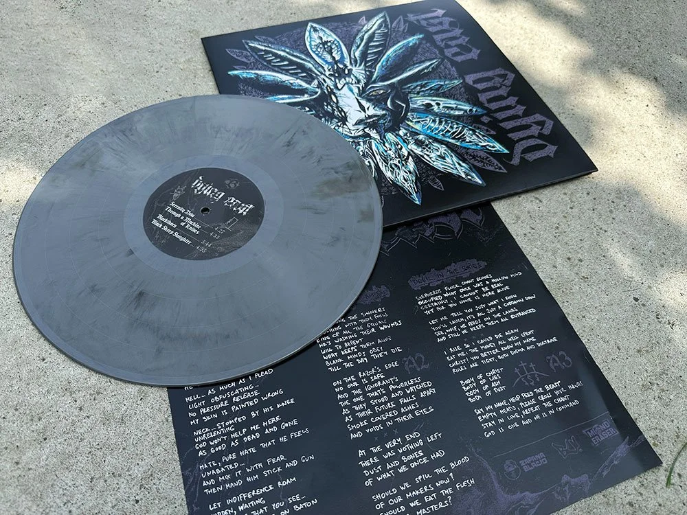 A vinyl record with a gray marble-like pattern and a black label, resting on and partly covering printed lyric sheets. The lyric sheets are black with handwritten white text and artwork of a detailed bird with angelic wings in shades of blue, white, 