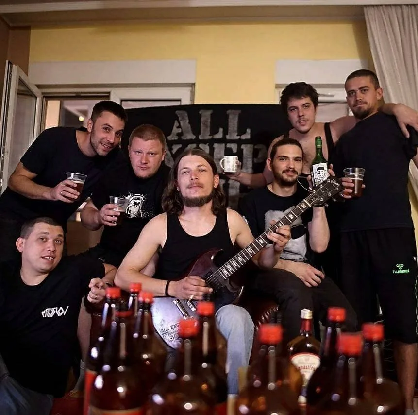 Group of seven young men celebrating with drinks, one playing a guitar, in a casual indoor setting with a yellow wall and curtains in the background.