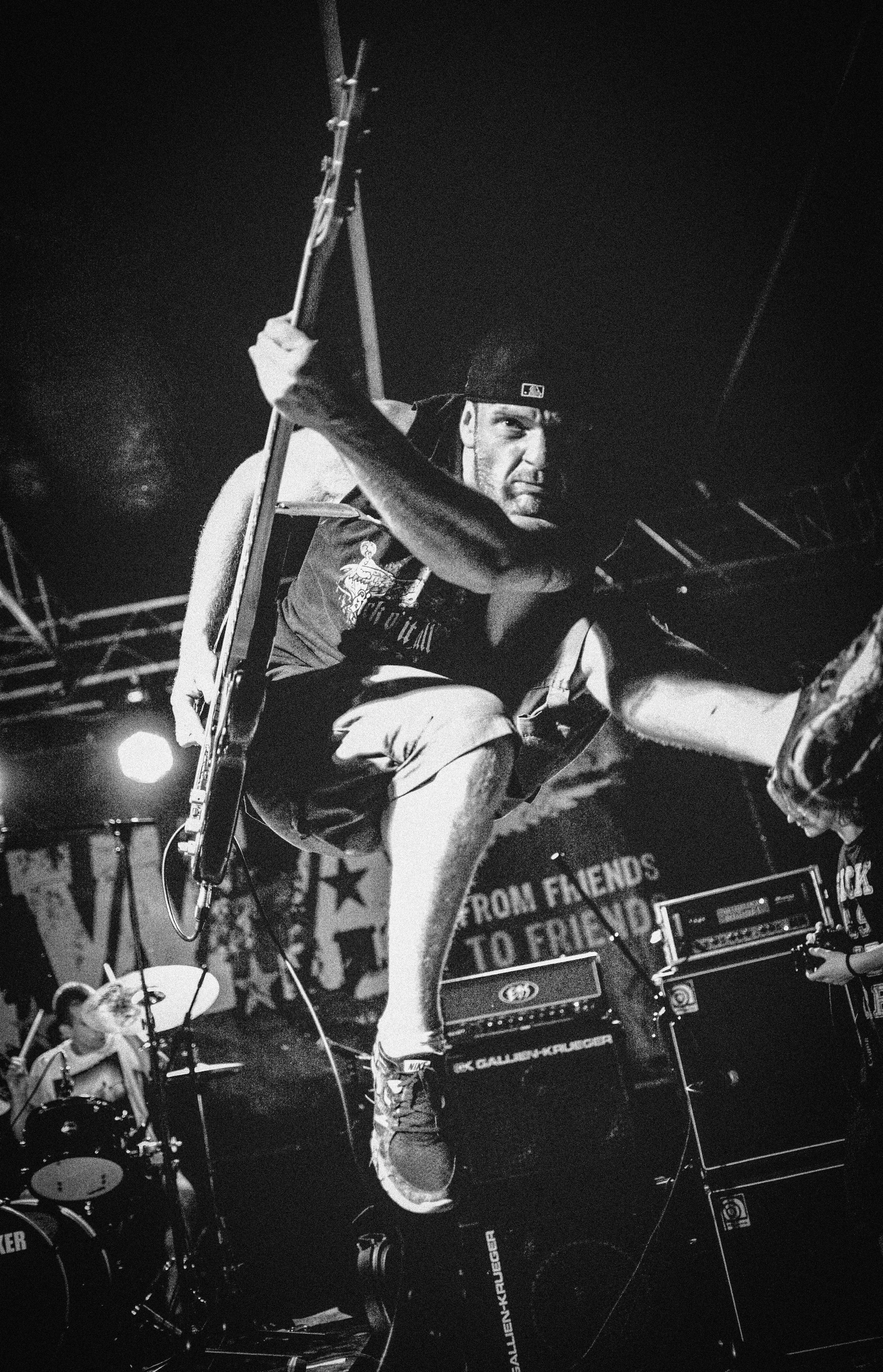 A male musician crouching on stage, holding a microphone stand with his left hand, with a serious expression, wearing a cap backwards and casual clothes, surrounded by musical equipment and drums, during a live performance.