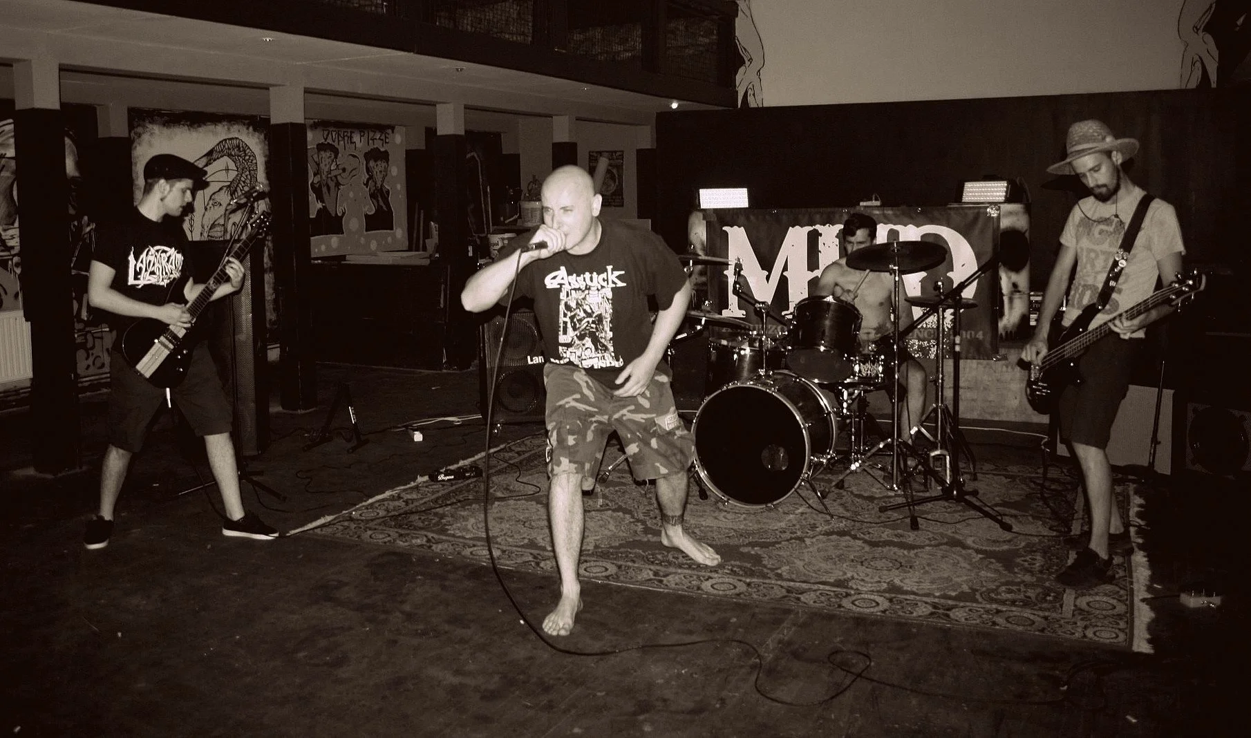 A band performing live on stage with four members: a vocalist holding a microphone, two guitarists, and a drummer. The vocalist is barefoot, wearing camouflage shorts and a black T-shirt. The stage has a rug, and there are artworks and posters on the