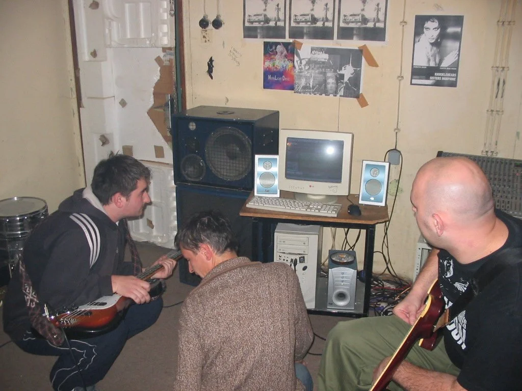 Three men in a room with music equipment, one playing guitar, one holding a guitar, and one kneeling, listening. The room has posters on the wall and a computer with speakers.