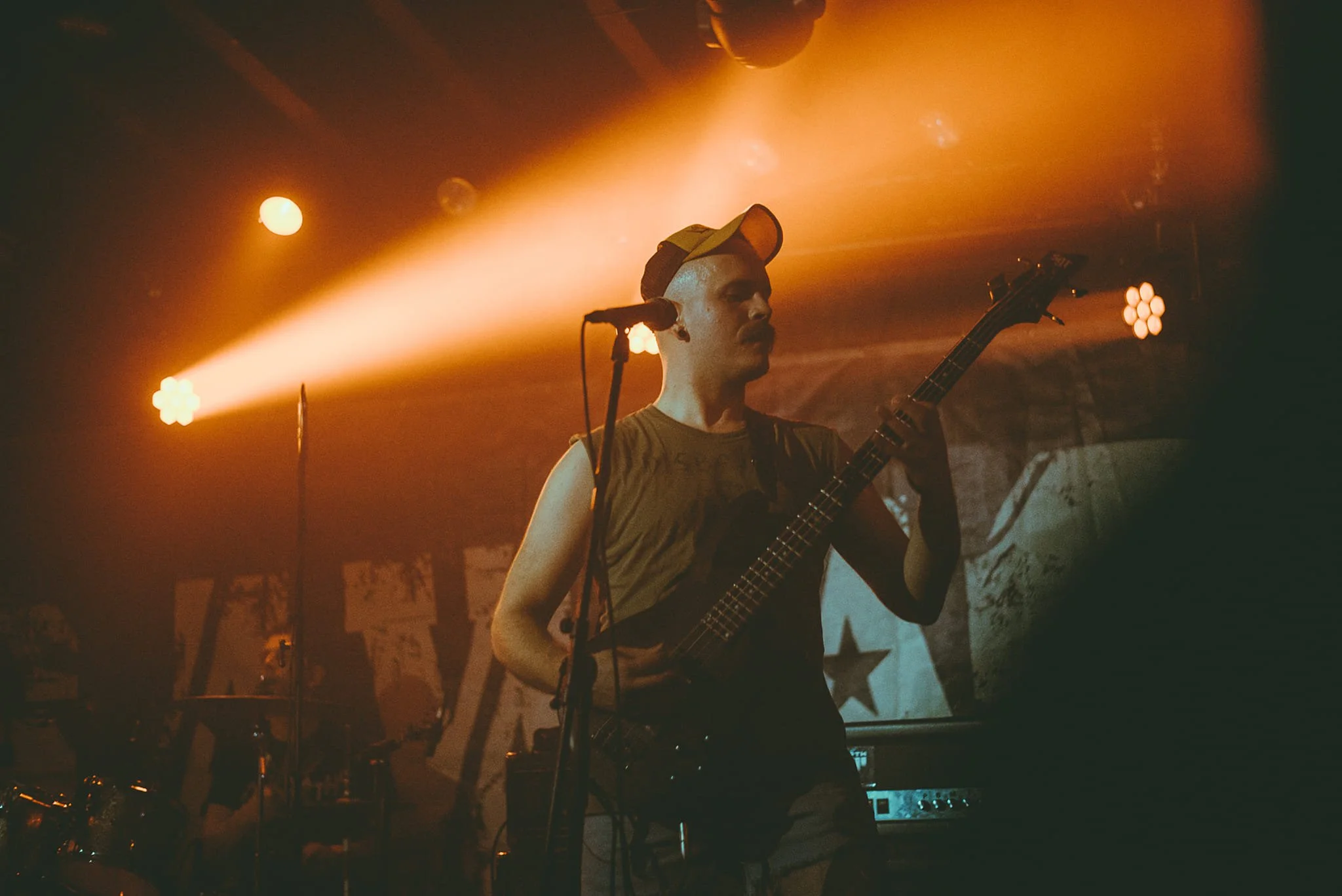 A male musician wearing a cap and sleeveless shirt playing an electric bass guitar on stage with orange lighting and microphones around him.