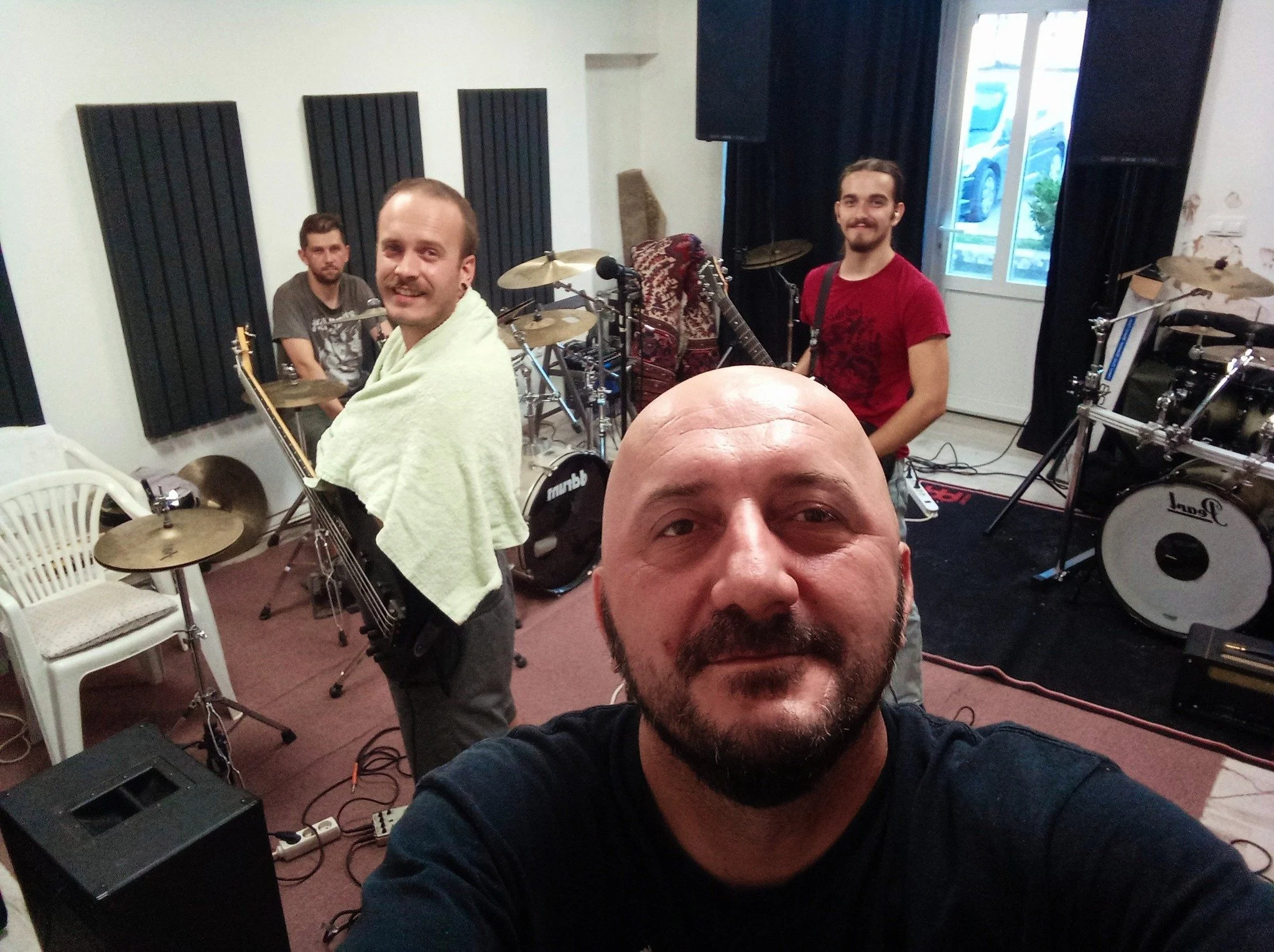 Four men in a music rehearsal room with drums and guitars, one man in the foreground taking a selfie, three men in the background looking at the camera, one wearing a towel over his shoulders.