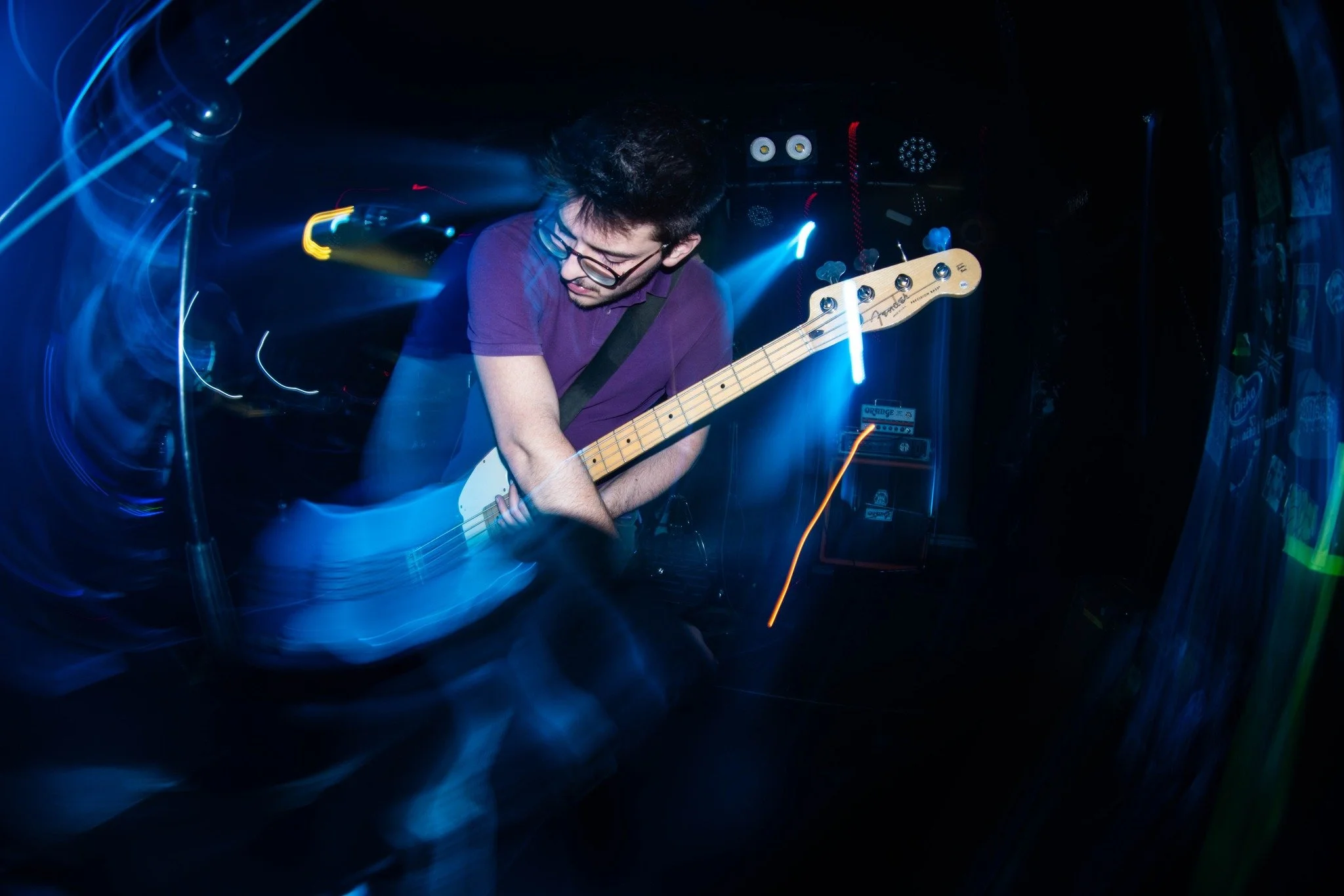 A young man with glasses playing an electric guitar on stage, surrounded by colorful lights and sound equipment, with motion blur creating a dynamic scene.