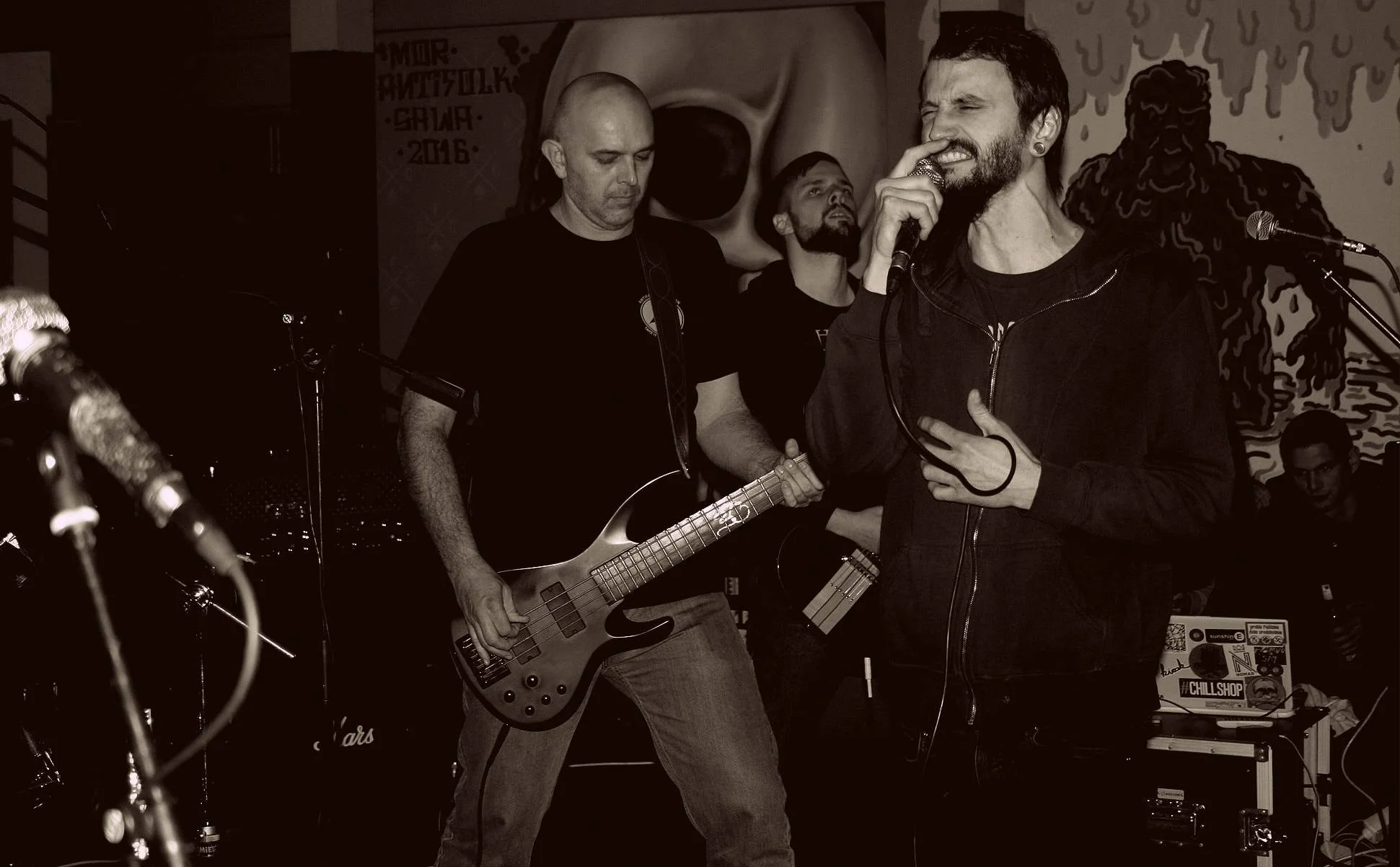 A band performing live on stage with a vocalist holding a microphone, a guitarist playing an electric bass, and other band members visible. The background has abstract art and posters.