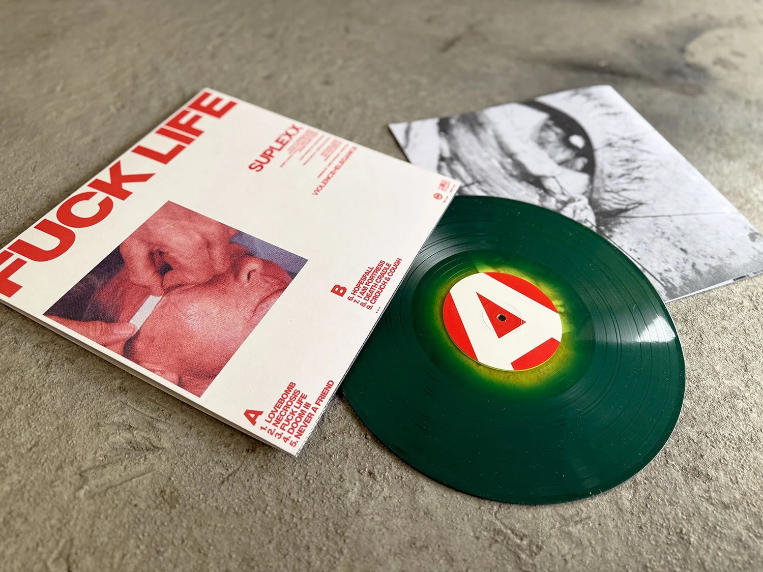 A vinyl record with a red and white label is partially out of its cover. The cover features a black-and-white photo and a tracklist in red text, with the title "FUCK LIFE" in large red letters.