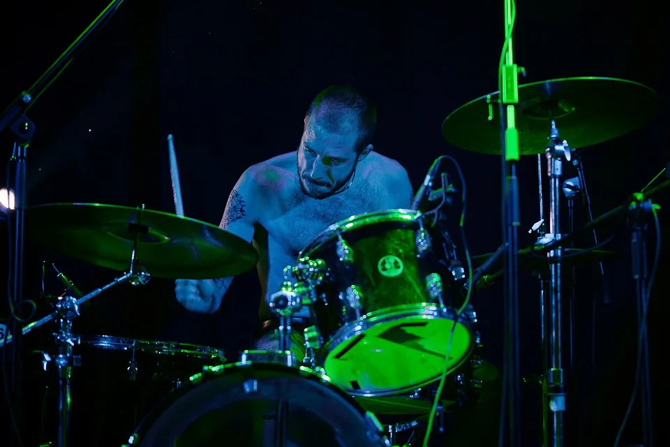 A shirtless male drummer playing a green drum set on stage, with dark background and stage lighting.