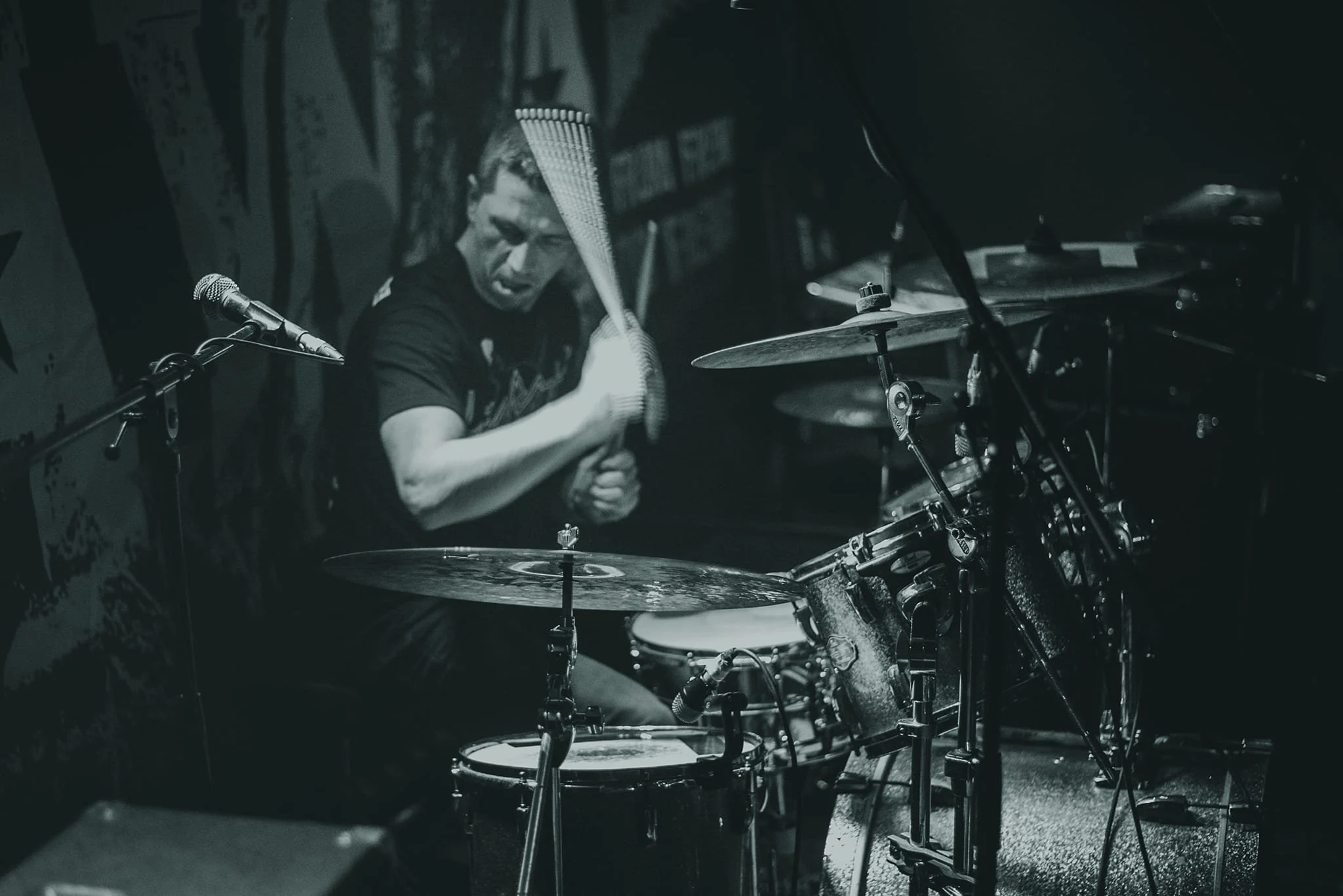A person playing the drums in a dimly lit room, with microphones set up around the drum kit, black and white photo.