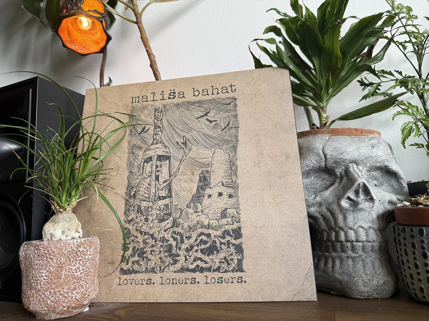 A framed artwork titled 'malisa bahat' with a sketch of a woman with long hair, a chain with a lock, and a landscape with a tower, surrounded by green potted plants on a wooden surface.
