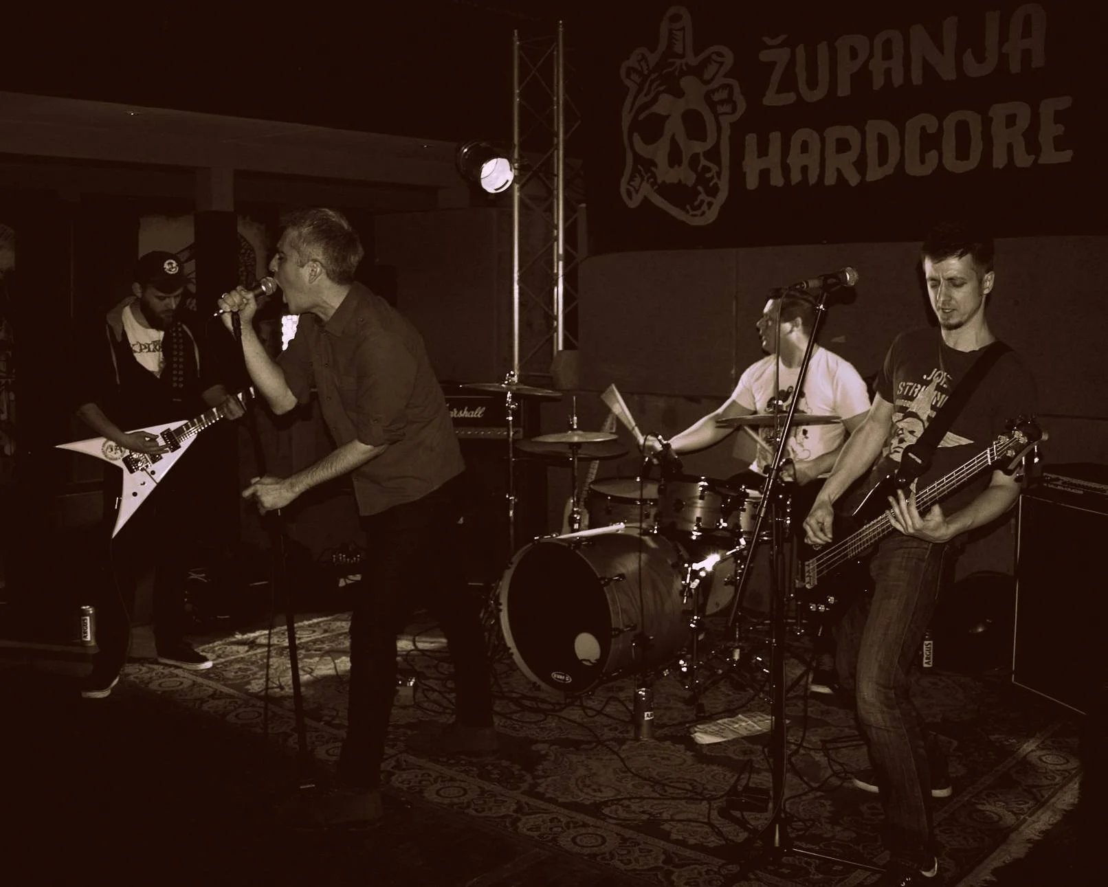 Four musicians performing on stage in a music venue, with a banner in the background reading 'Župa Hardcore'. One person is singing into a microphone, another is playing an electric guitar, a third is playing a drum set, and the fourth is playing a b