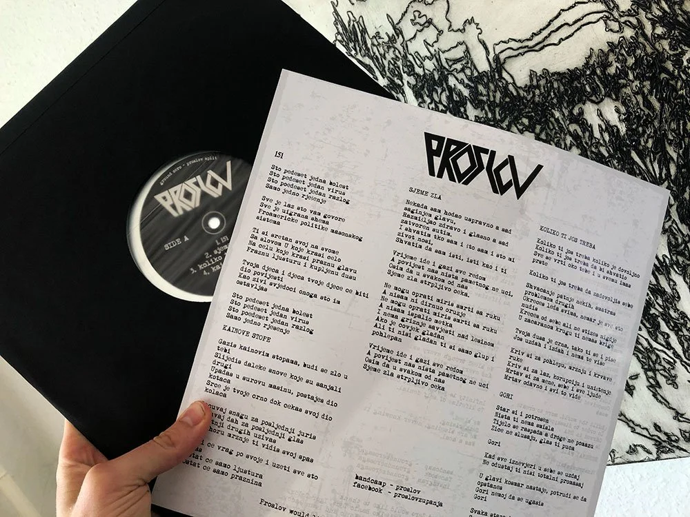 A black vinyl record partially pulled out of its sleeve, revealing a label with the word PROSLOV. There is also a printed sheet with lyrics or text in a Slavic language, and an illustration of trees and nature in the background.